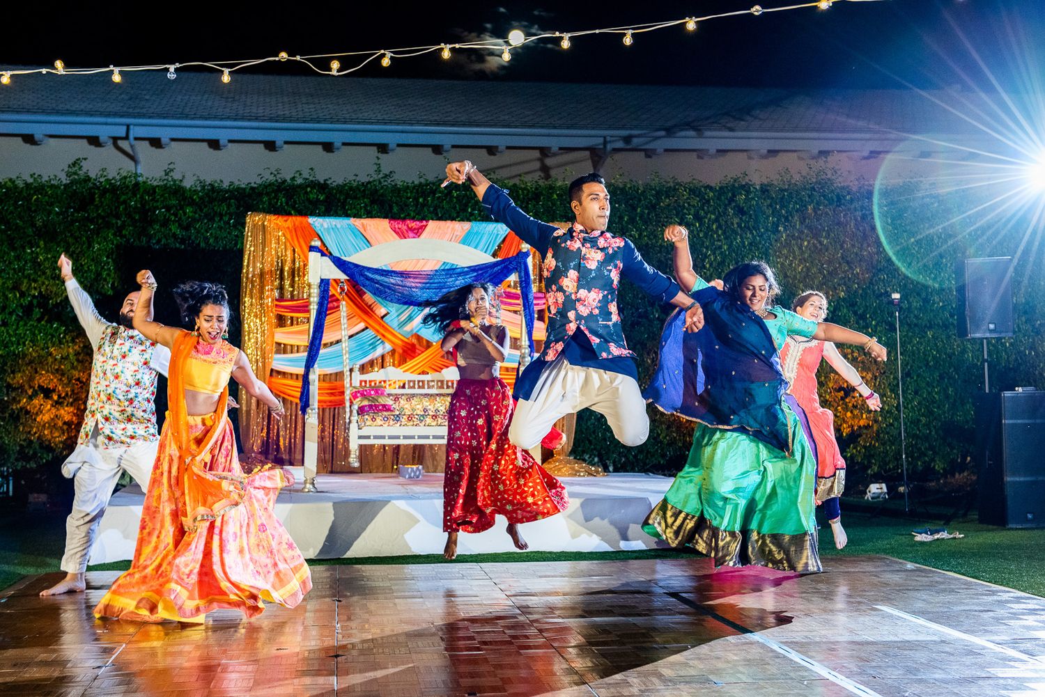 Colorful outdoor wedding performance with dancers in traditional Indian outfits under string lights.