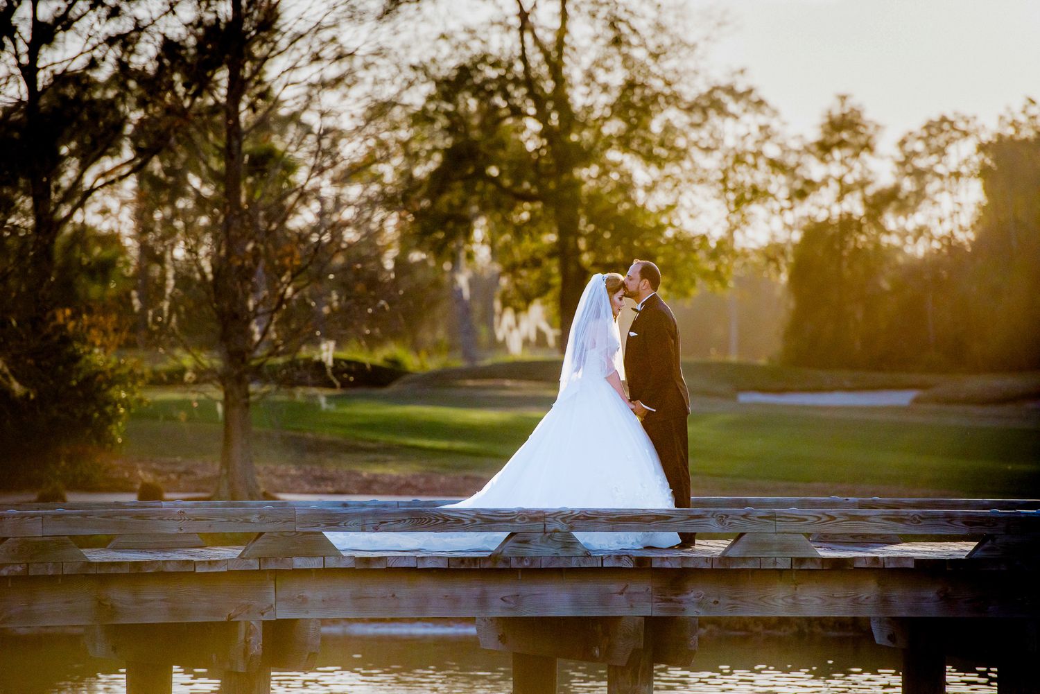 Silhouetted couple stands on wooden dock at sunset creating romantic wedding portrait against golden sky.