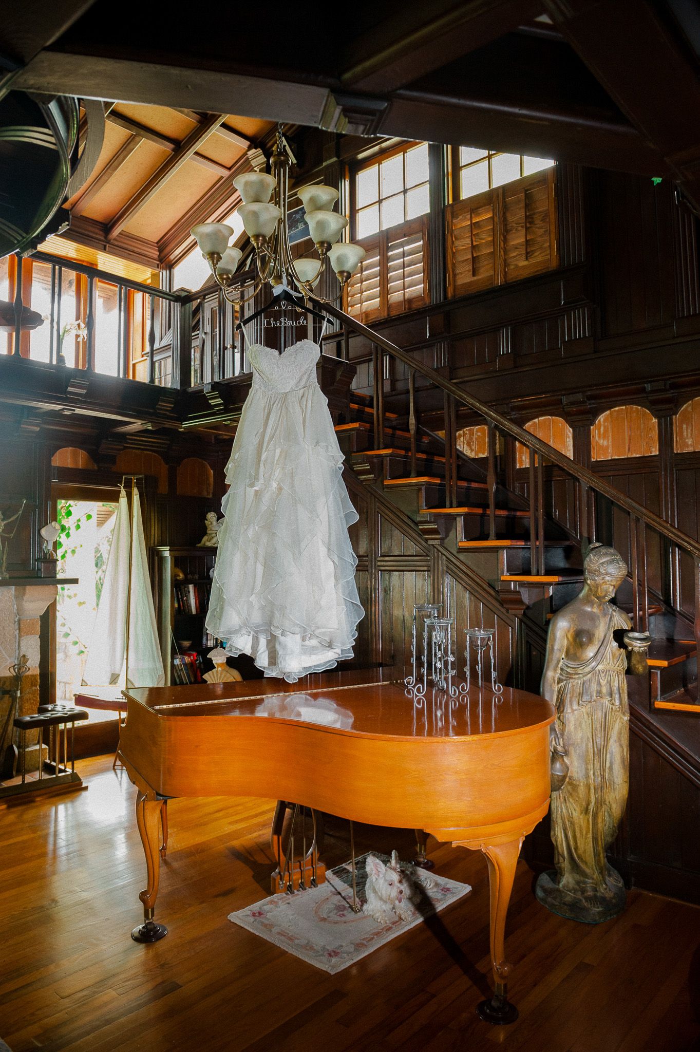 A white wedding dress hangs elegantly above a grand piano in a rustic wooden interior with natural lighting.