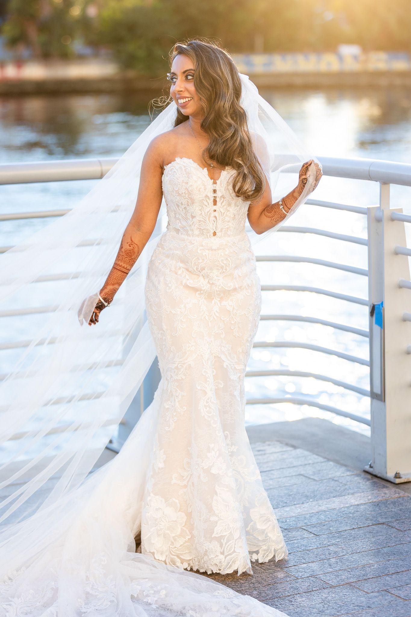 A bride in a fitted lace wedding gown poses on a waterfront deck at sunset.