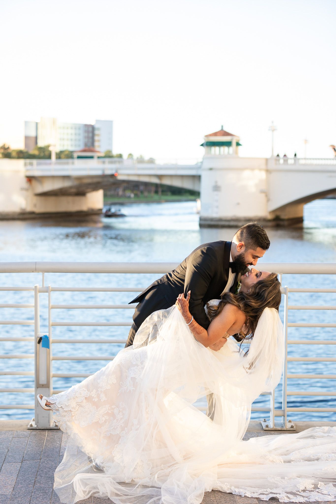 Romantic dip kiss on waterfront with flowing white dress and bridge in background.