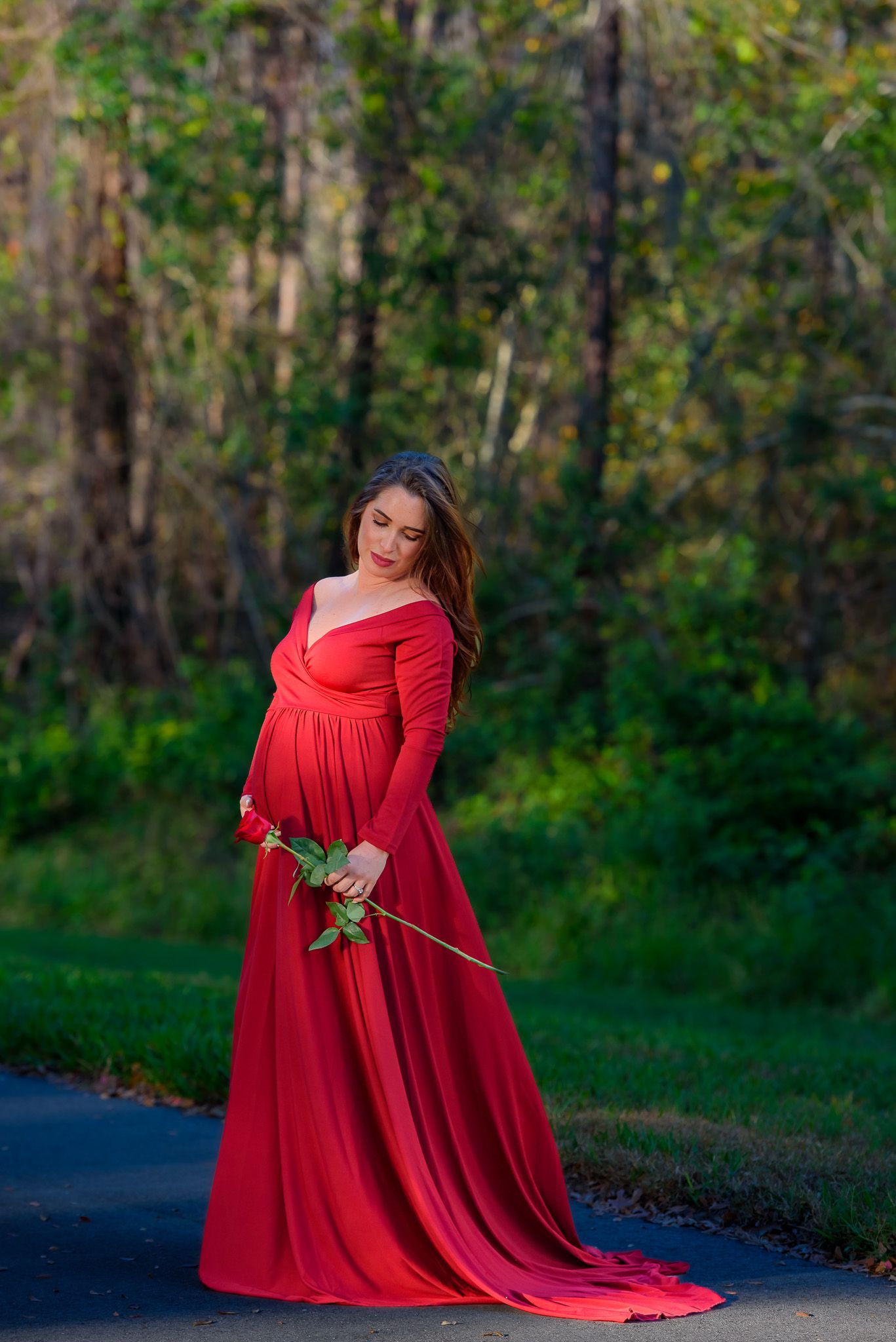 A flowing red maternity gown with an off-shoulder design photographed against a green forest background.