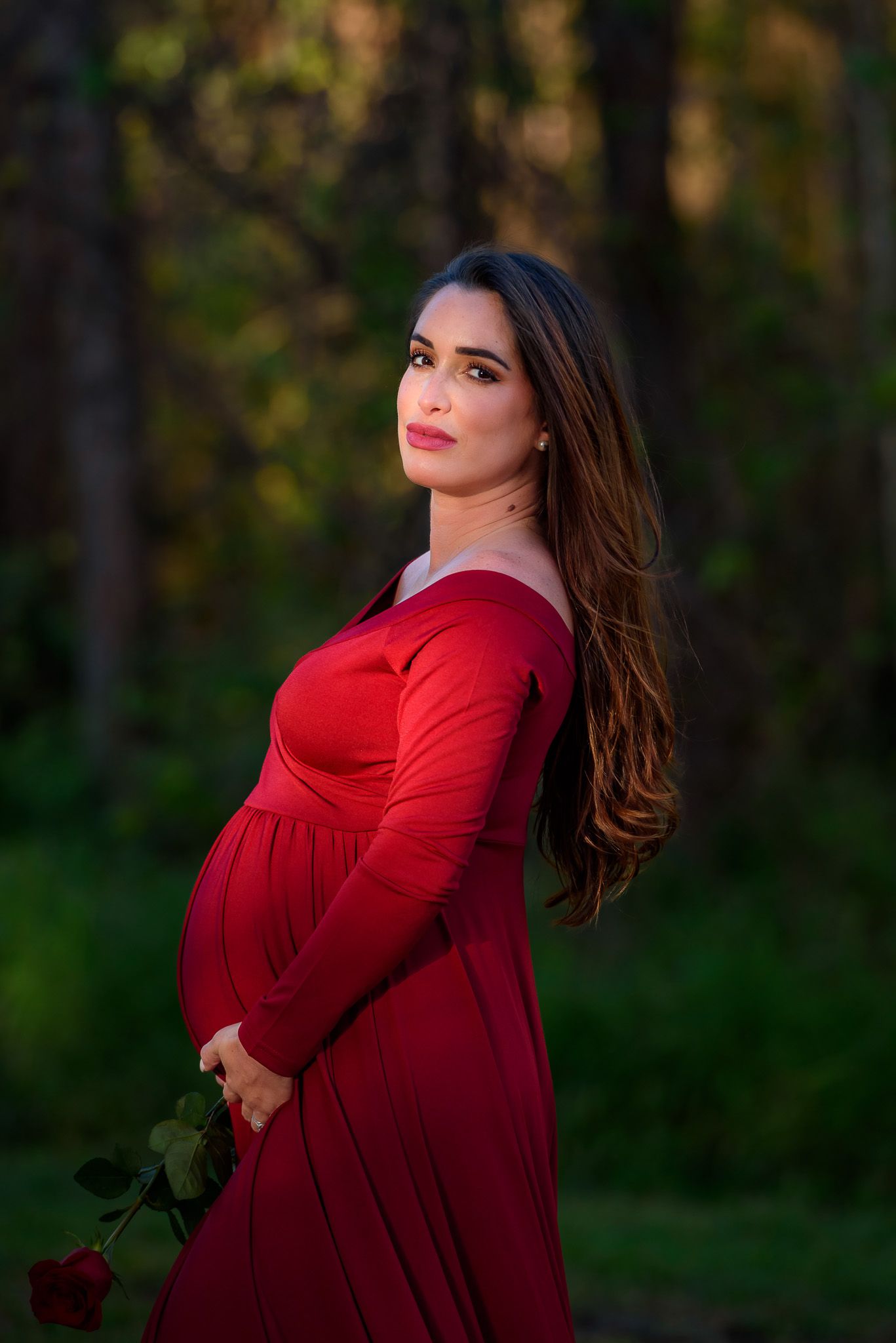 Maternity portrait of woman in red dress against forest backdrop at golden hour.