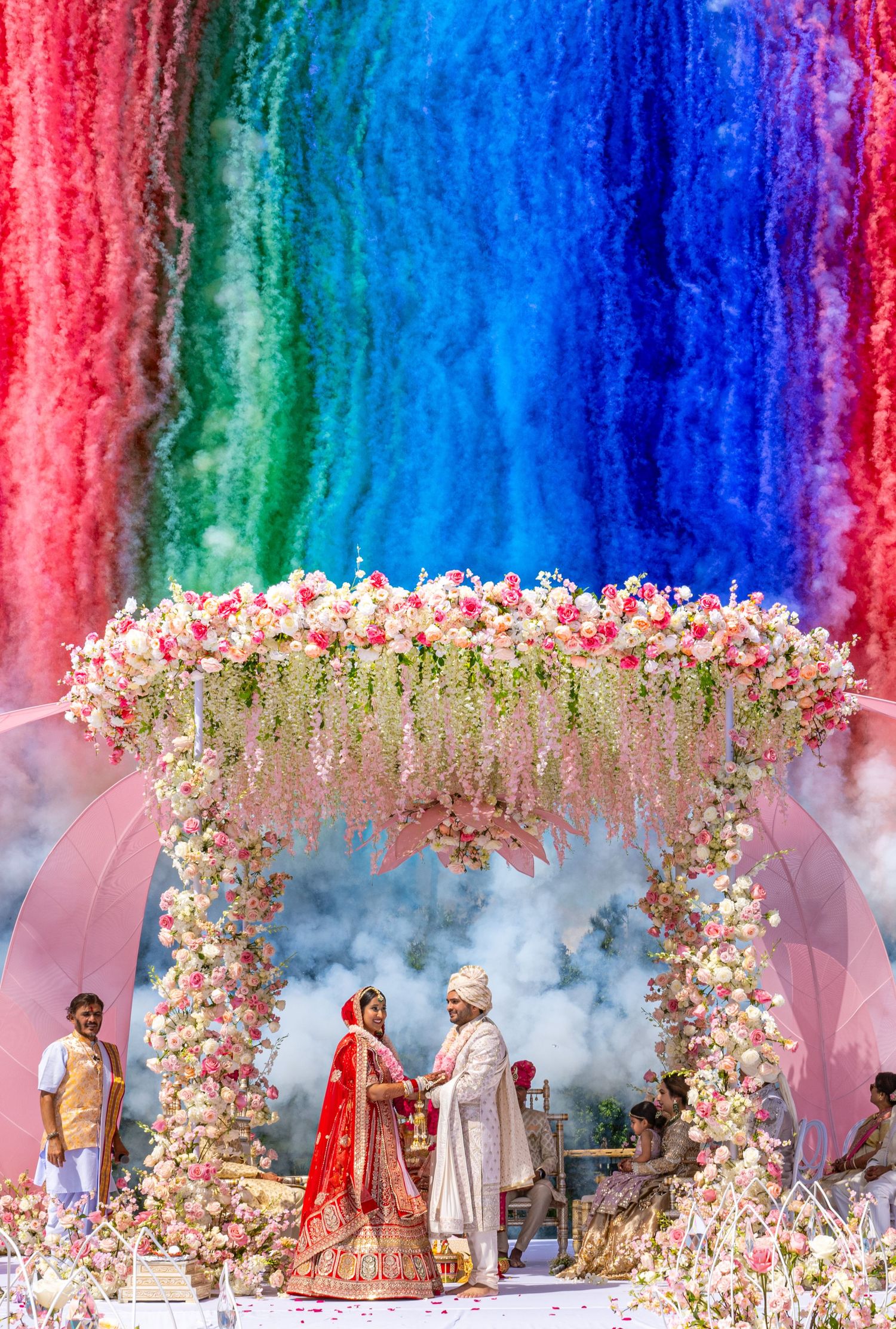 Colorful wedding ceremony under a floral arch with rainbow backdrop and pink flower decorations.