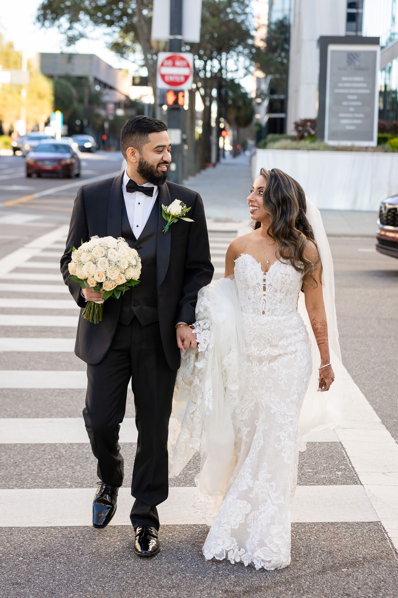 A wedding couple walking across a crosswalk with the bride holding a bouquet of white roses.