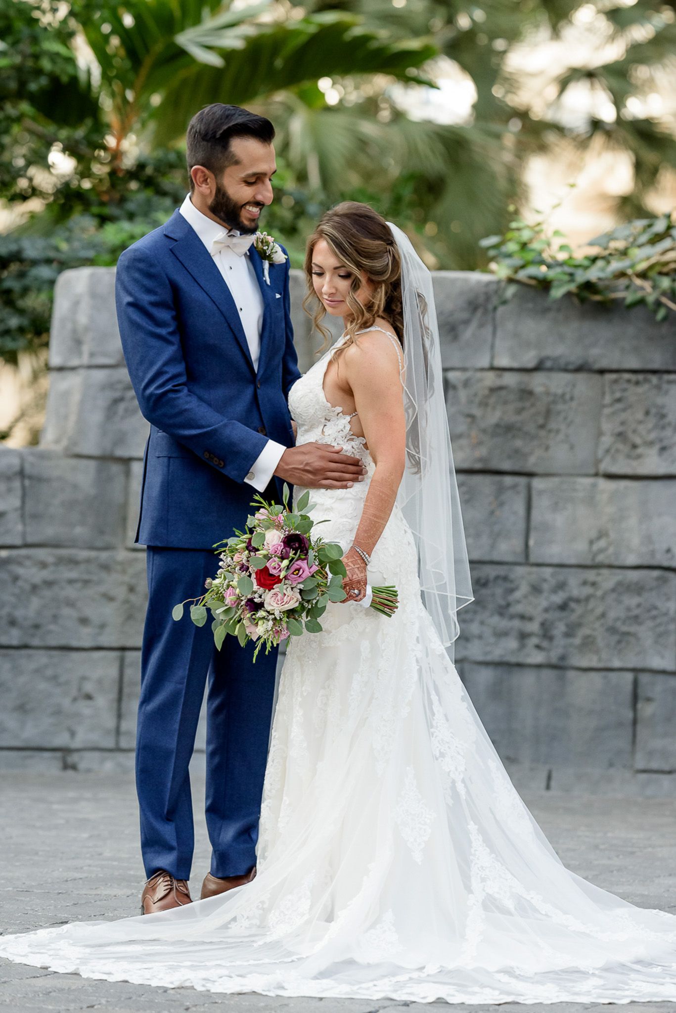 Couple in formal wedding attire posing with floral bouquet against stone wall.