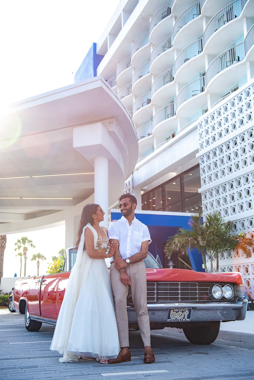 A couple poses beside a vintage red car in front of a retro-modern hotel with white architecture.