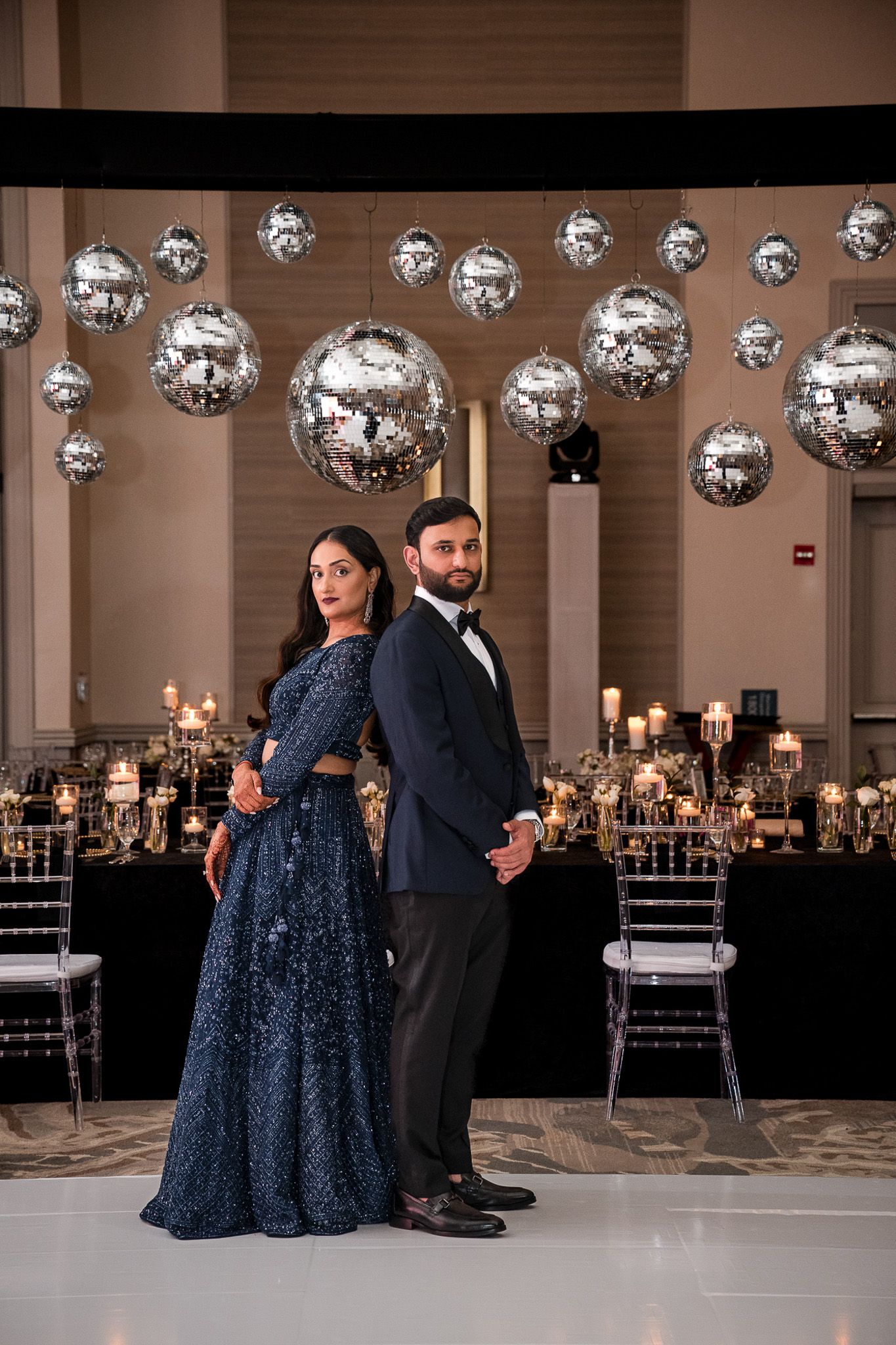Elegant couple poses back-to-back under silver disco balls in a luxurious reception setting.