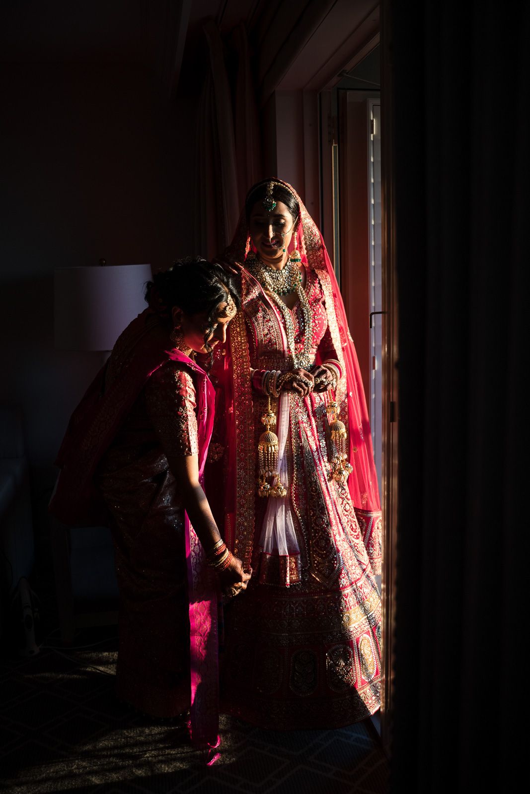 Dramatic lighting illuminates a bride in red traditional lehenga as her dress is adjusted.