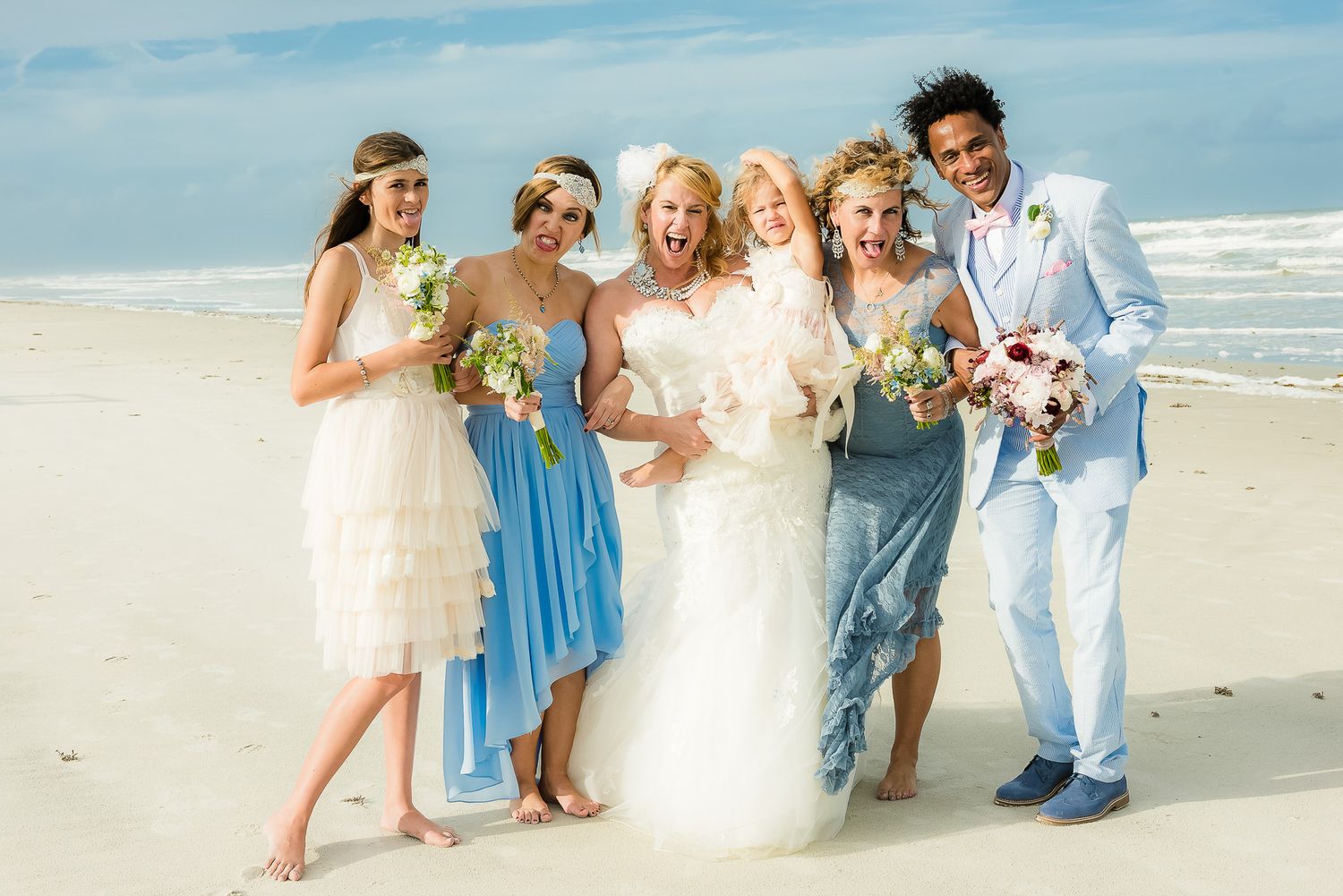 Wedding party in mix of blue and white attire poses joyfully on sunny beach.
