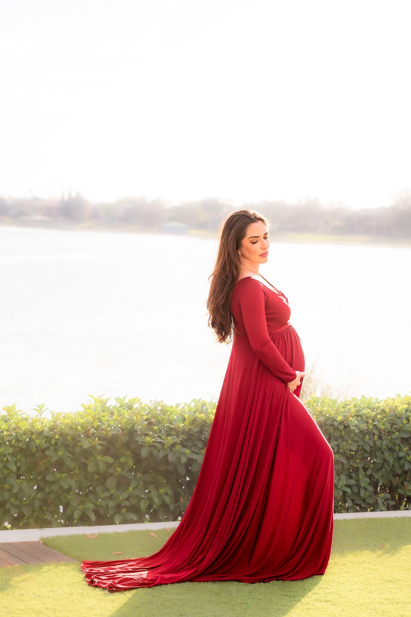 Pregnant woman in flowing red dress poses at sunset near waterfront.