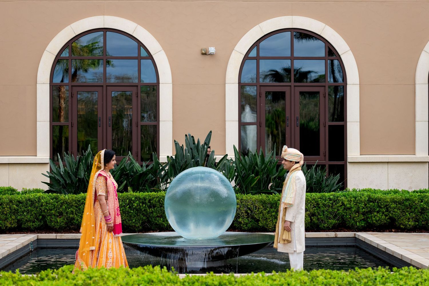 Two people in traditional attire stand by a spherical water feature in front of arched windows.