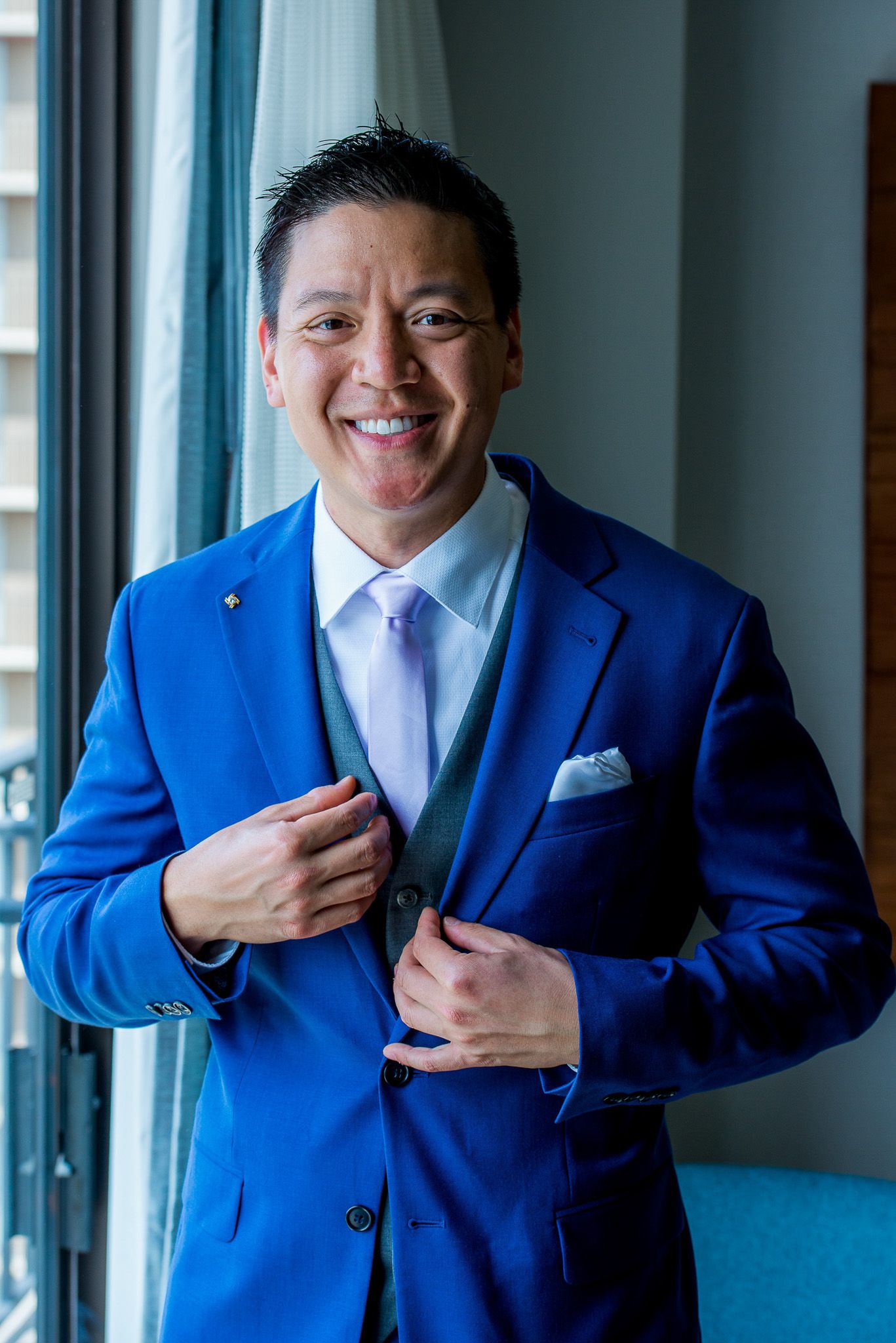 Professional wearing a vibrant royal blue suit jacket and light tie smiles warmly by a window.