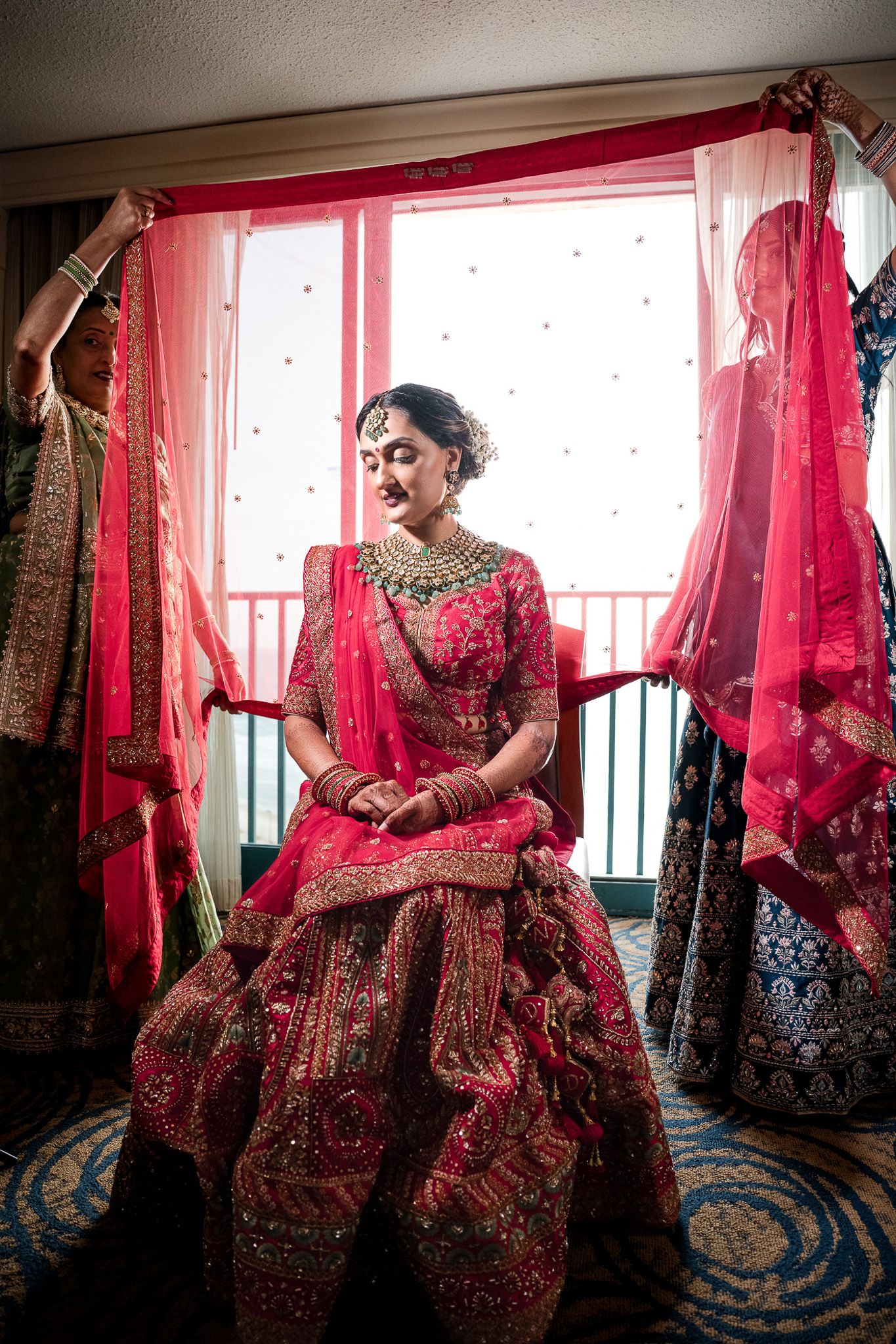 Traditional South Asian bride in red lehenga poses by window with flowing drapery.