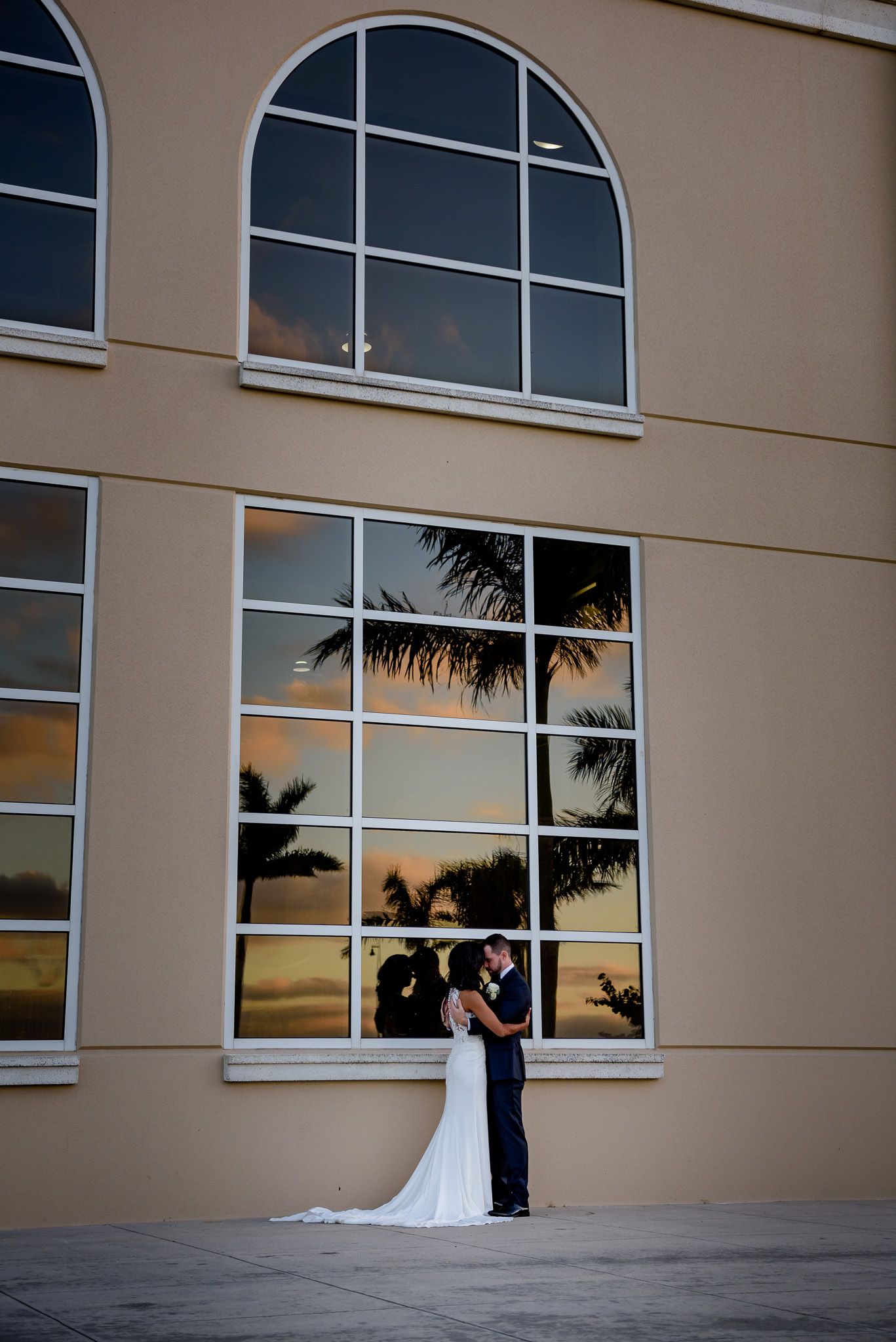 Couple shares intimate moment against large windowed building at sunset with palm tree reflections.