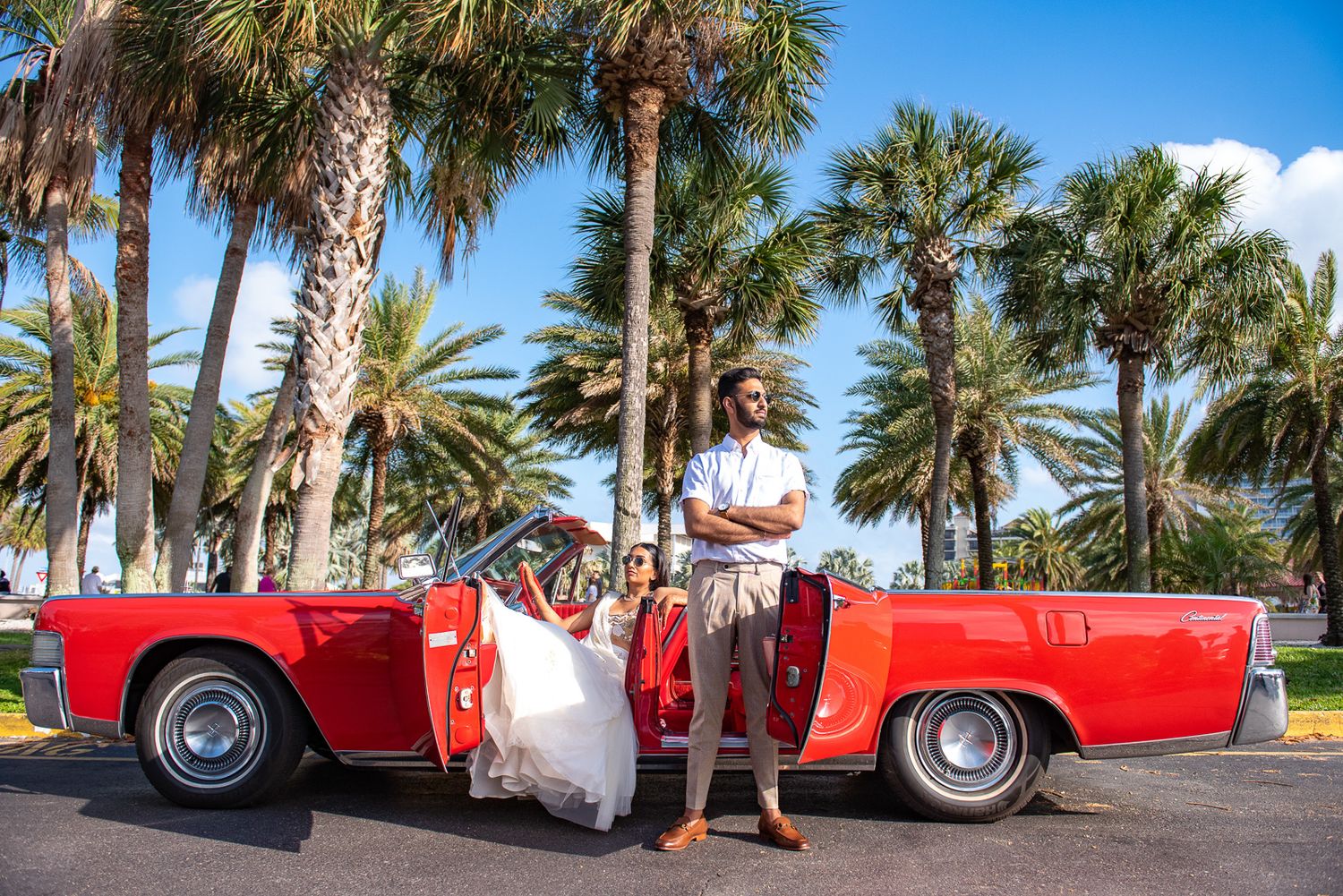 Newlyweds posing with vintage red convertible car among tropical palm trees.