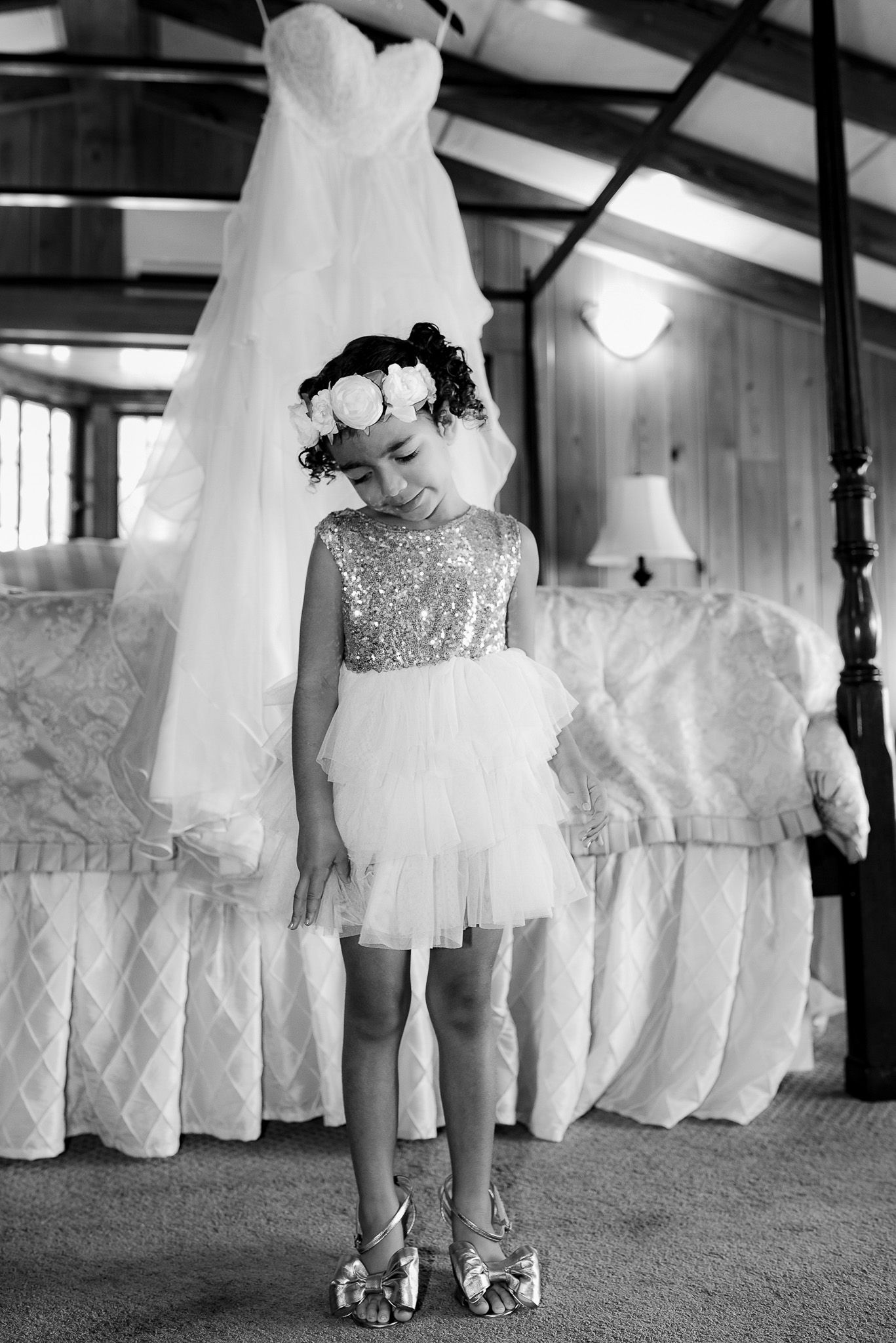 Black and white photo of flower girl in sparkly dress standing beside wedding gown.