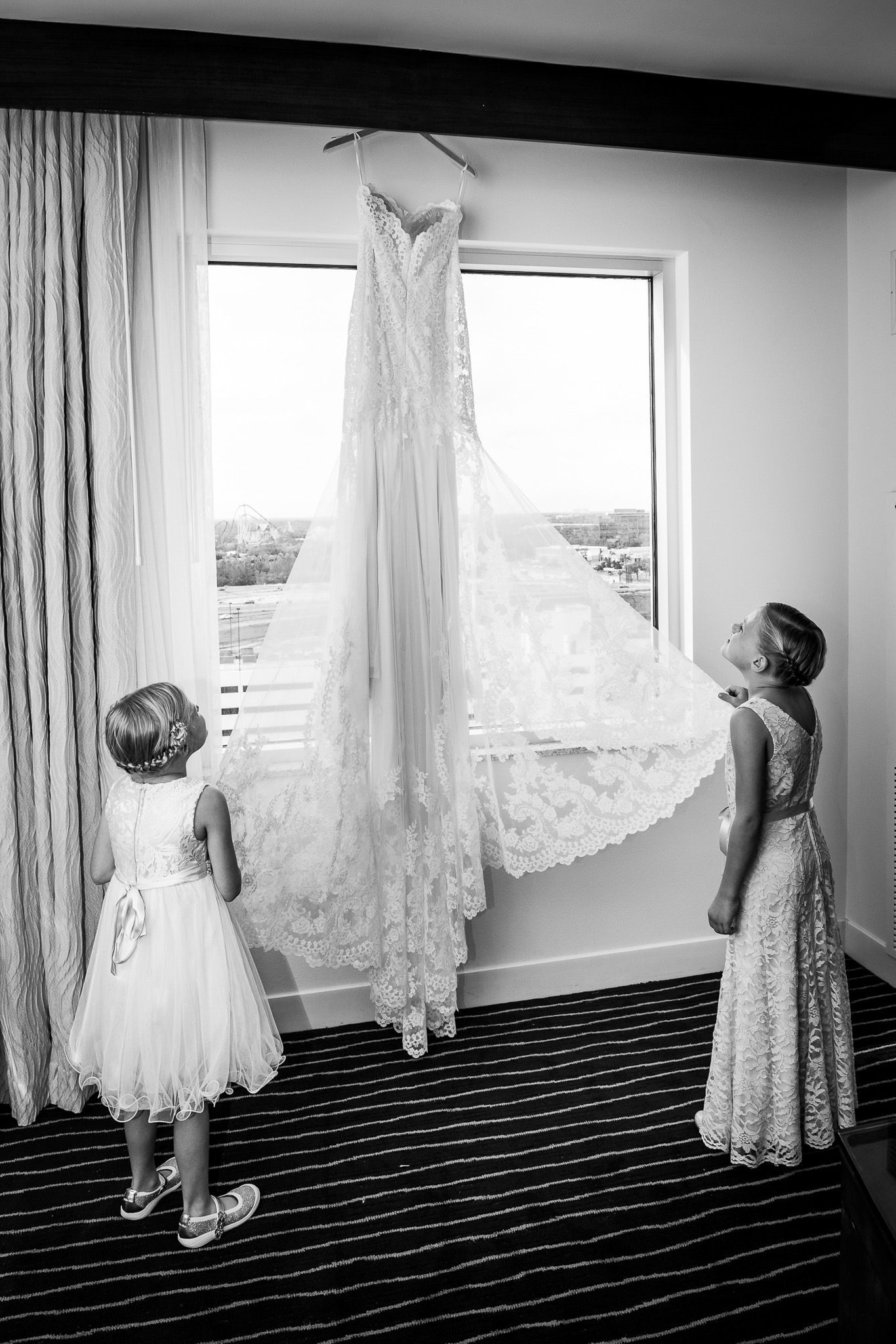 Two people admiring an elegant white lace wedding dress suspended in front of a window with natural light.