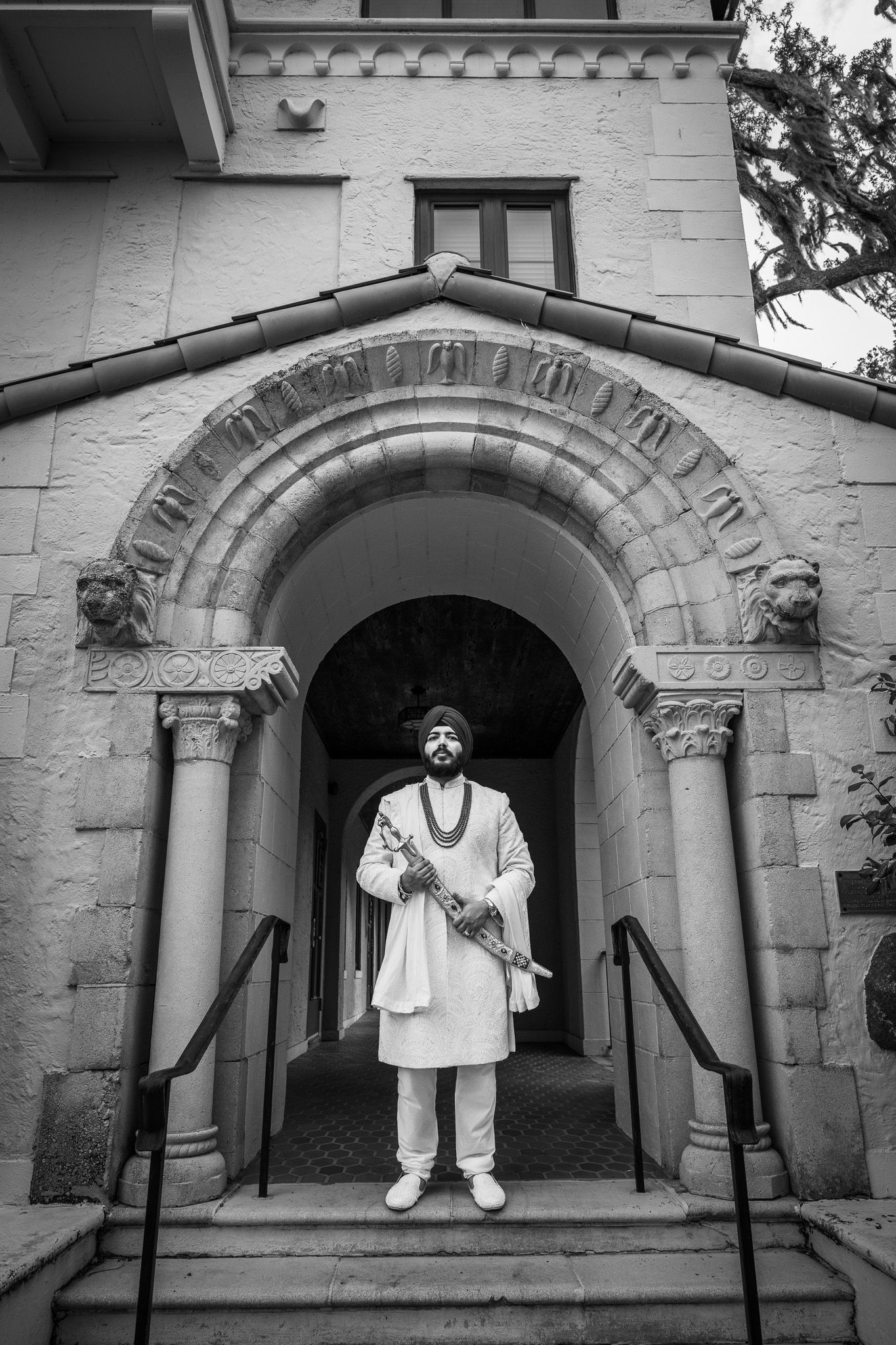 Figure in traditional white attire stands beneath ornate stone archway entrance of historic building.