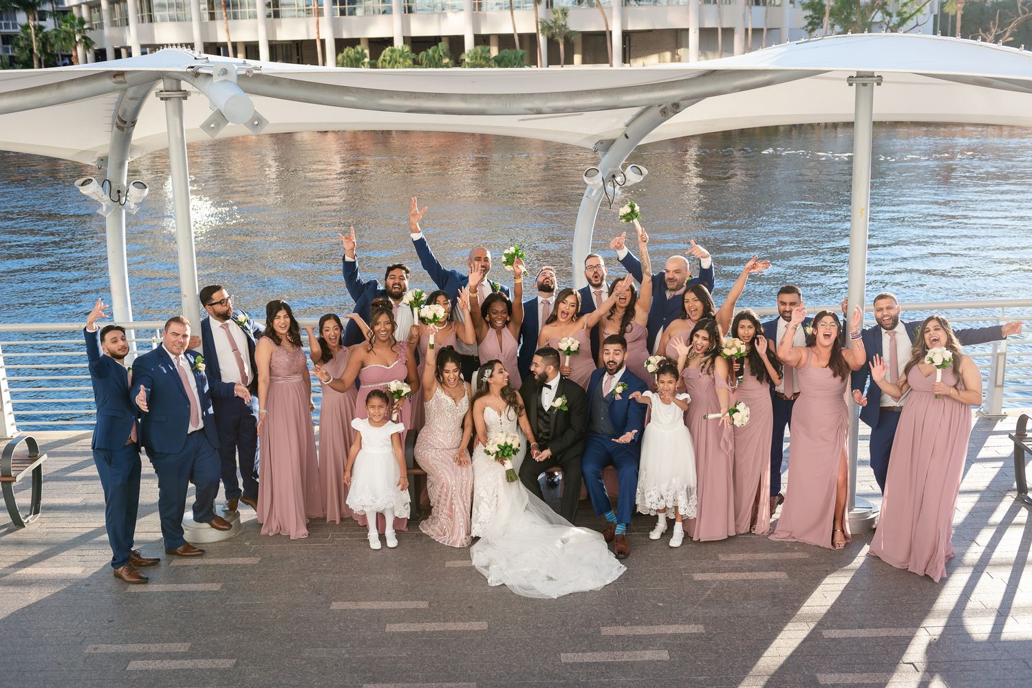 A large wedding party poses on a waterfront deck with everyone cheering.