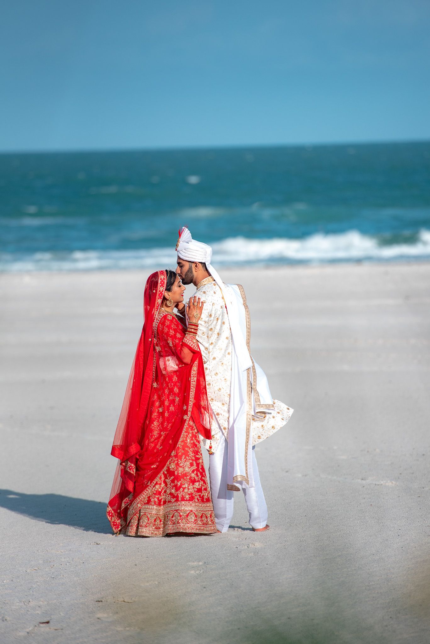 A couple in traditional wedding attire share an intimate moment on a sandy beach.