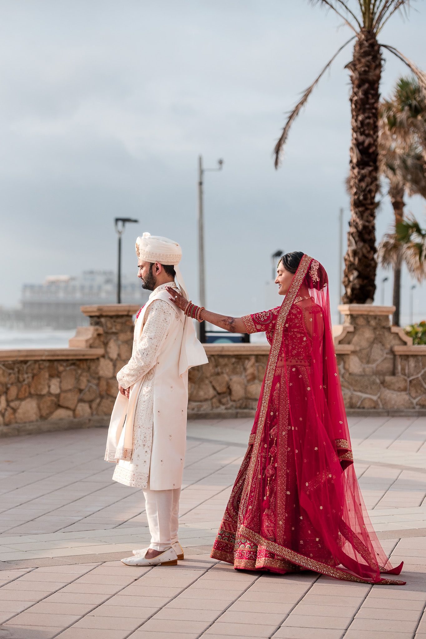 South Asian wedding couple in traditional attire sharing a moment on oceanfront promenade.