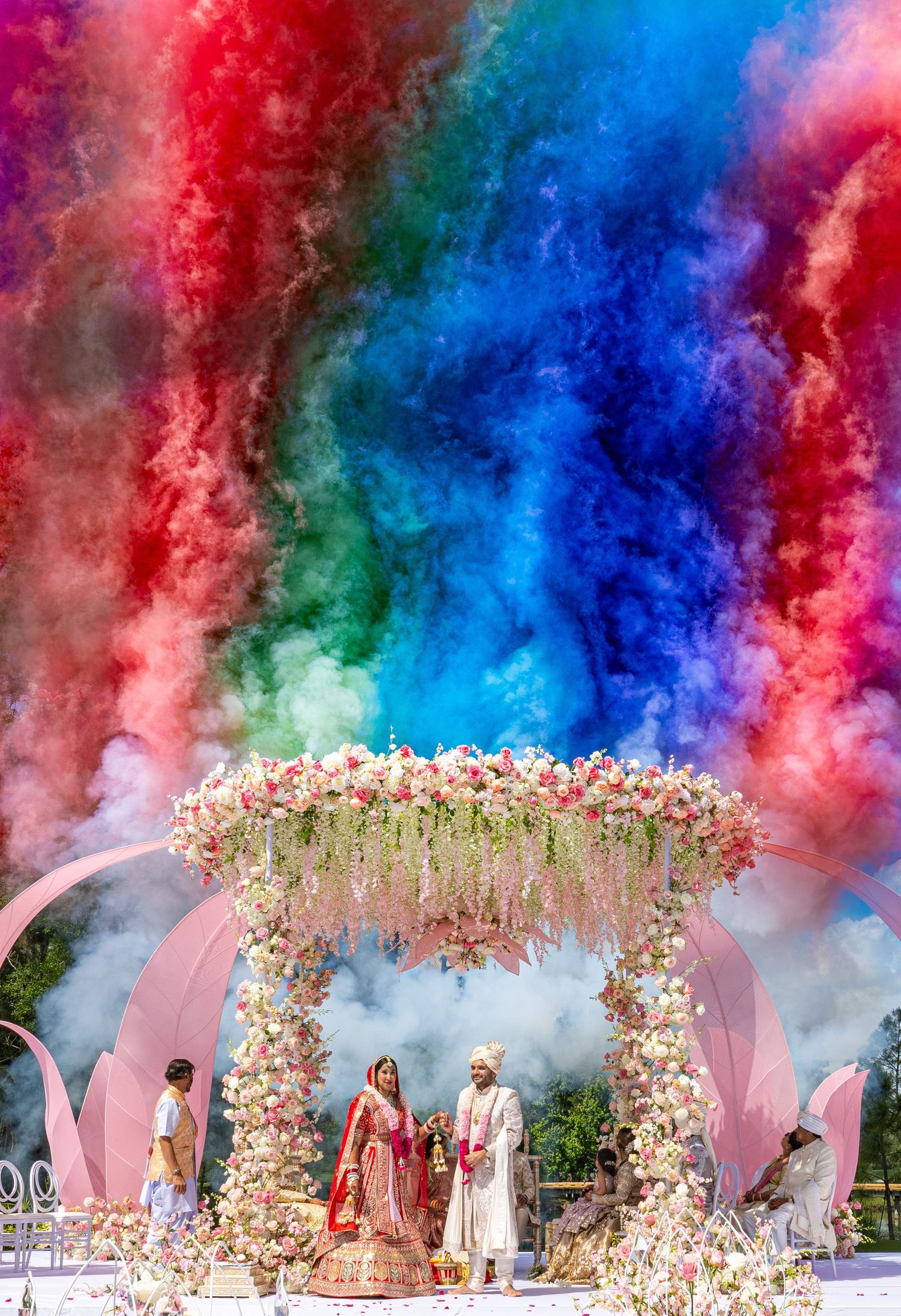 A floral wedding mandap with vibrant colored smoke effects creating a dramatic backdrop for the ceremony.