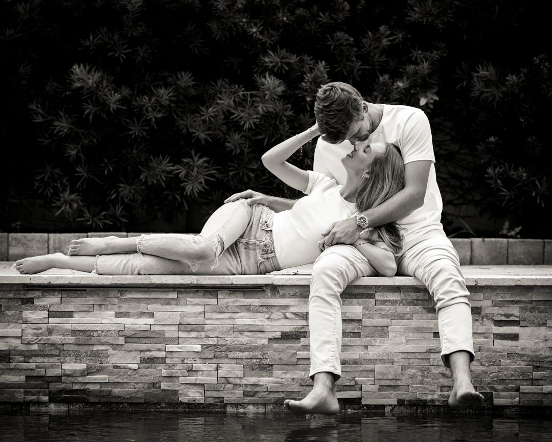 Intimate black and white photo of couple reclining on stone wall by water.