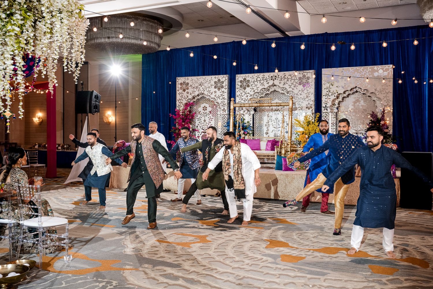 Group performing lively dance moves at wedding reception with blue backdrop and hanging lights.