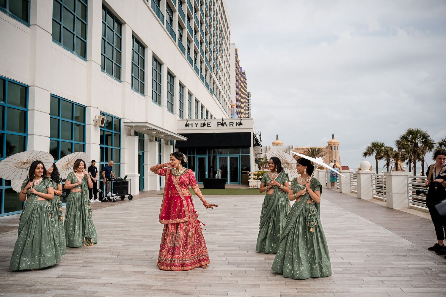 Bridesmaids in sage green dresses pose with bride in red lehenga on beachfront boardwalk.
