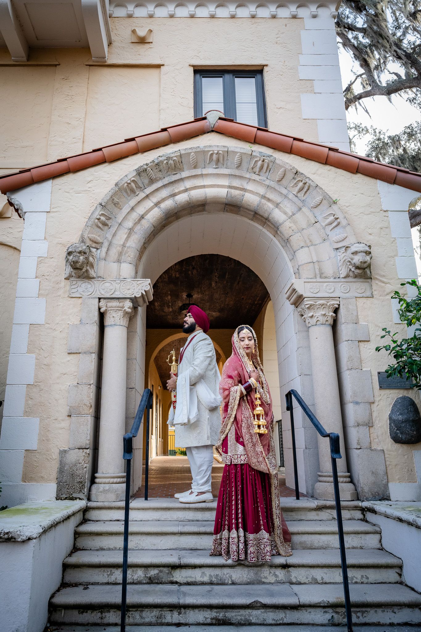 Couple in traditional Indian wedding attire pose on stone steps under arched entrance.