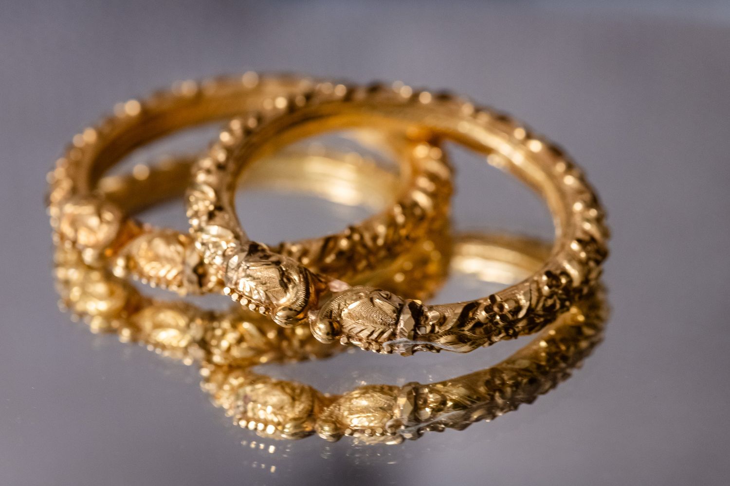 Close-up of ornate gold wedding rings with intricate designs reflecting on a mirrored surface.