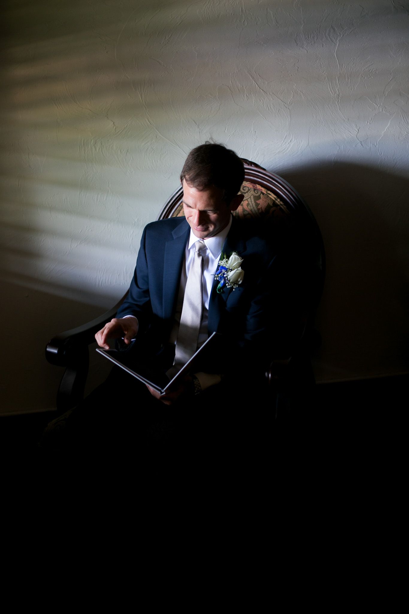 Dramatic lighting captures a groom reading in formal attire while seated in an antique chair.