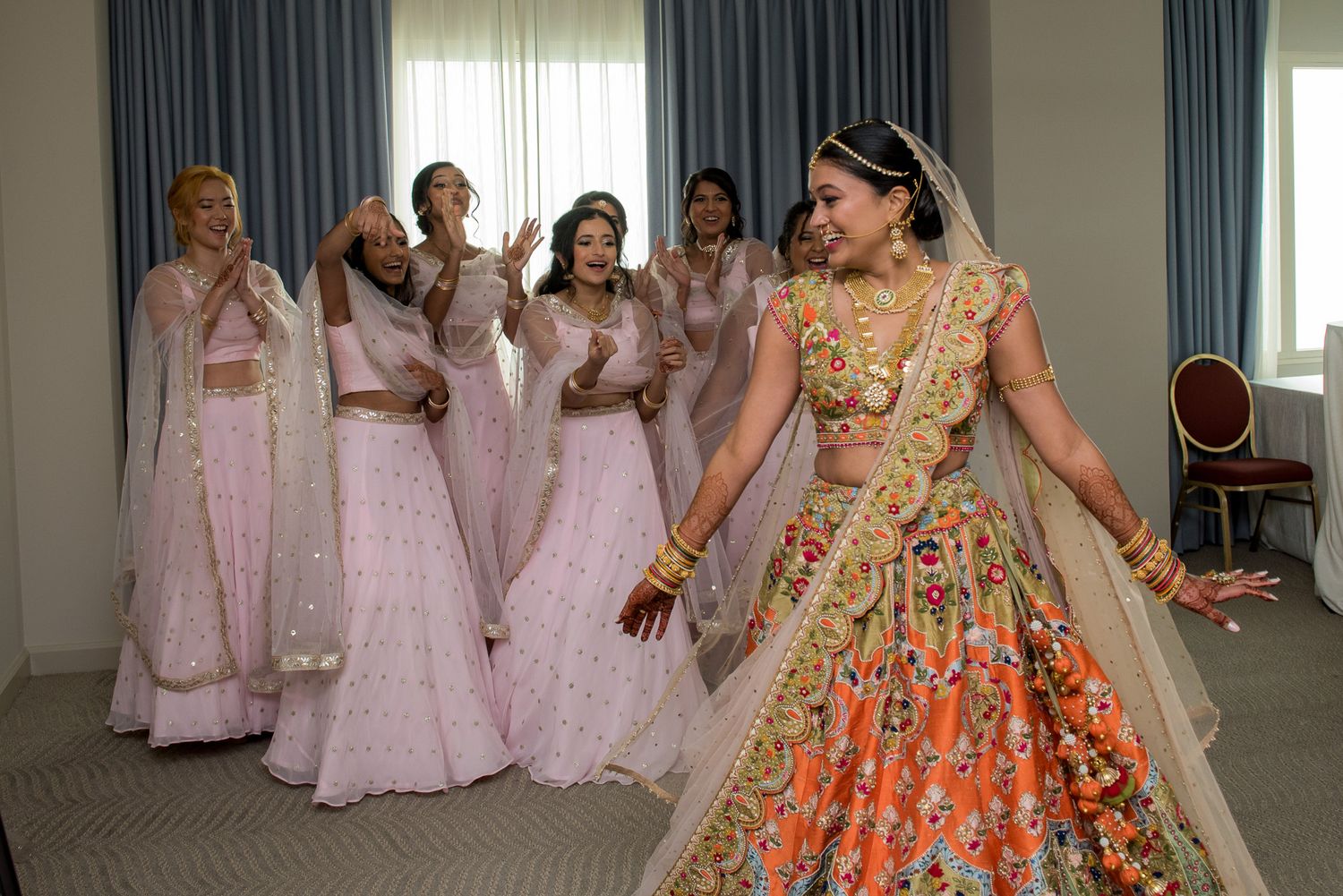 Bride in ornate orange lehenga celebrates with bridesmaids in pink dresses.