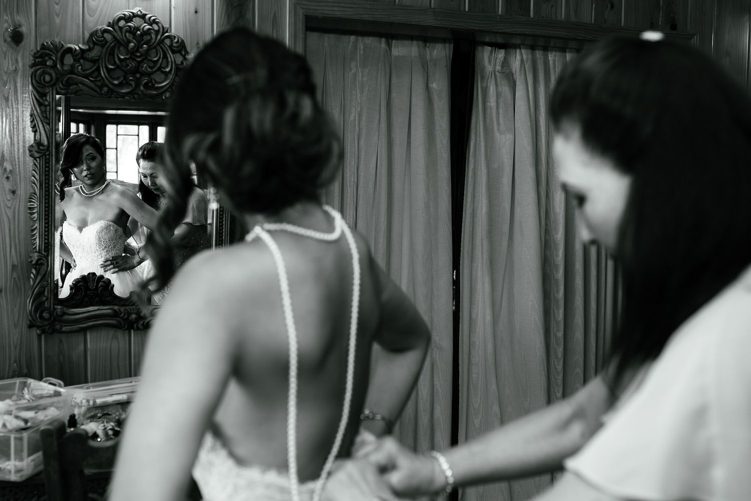 Black and white photo of wedding preparations in a room with a mirror and curtains.