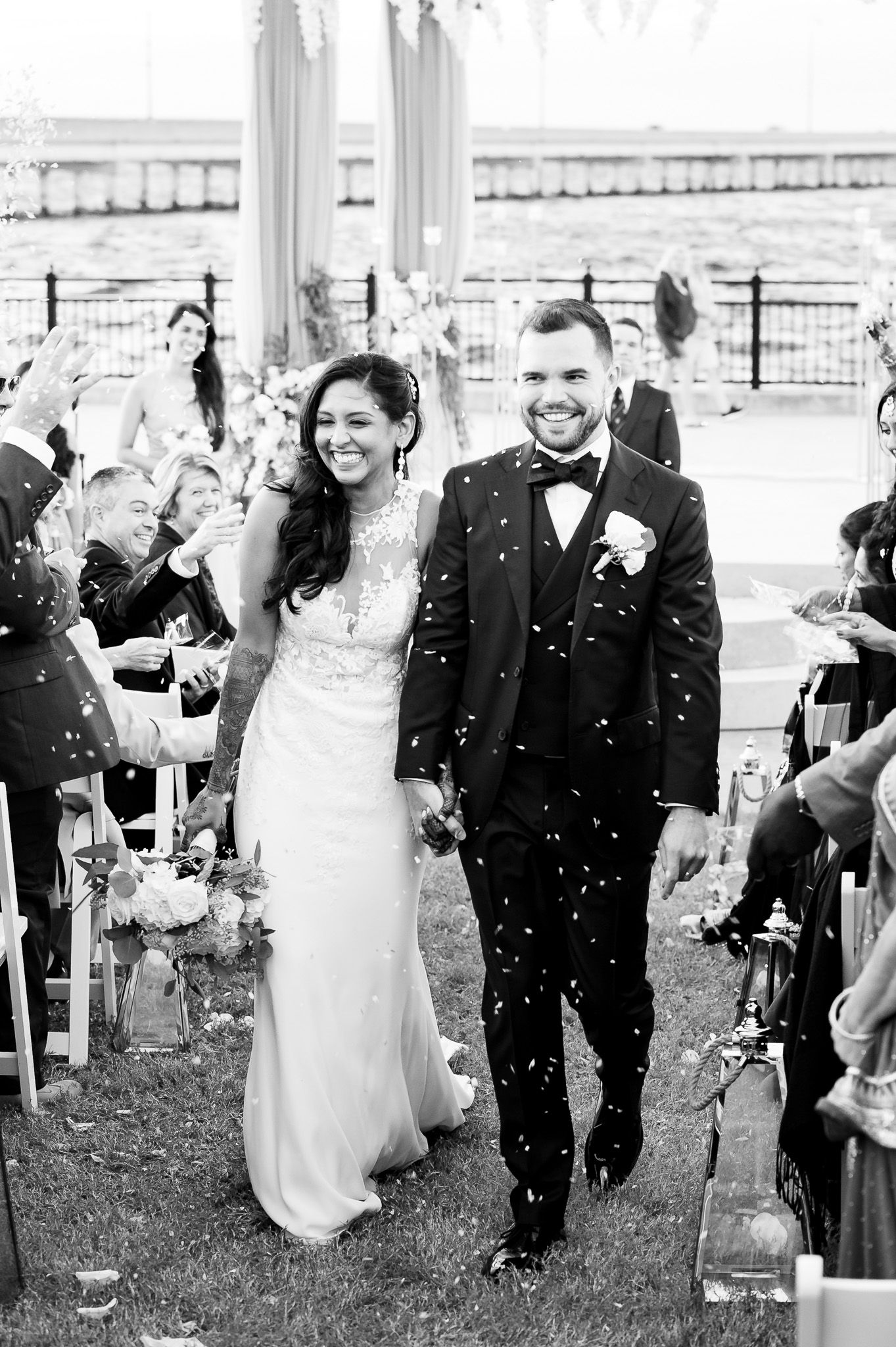 Wedding couple walks down aisle while guests throw confetti during outdoor waterfront ceremony.