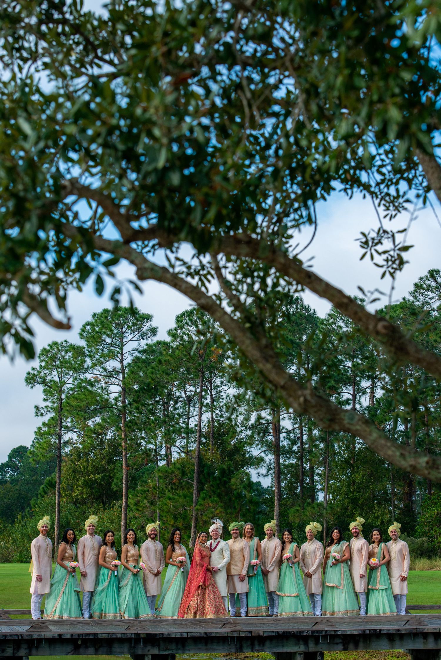 Wedding party in pastel outfits standing on wooden dock with tall pine trees in background.