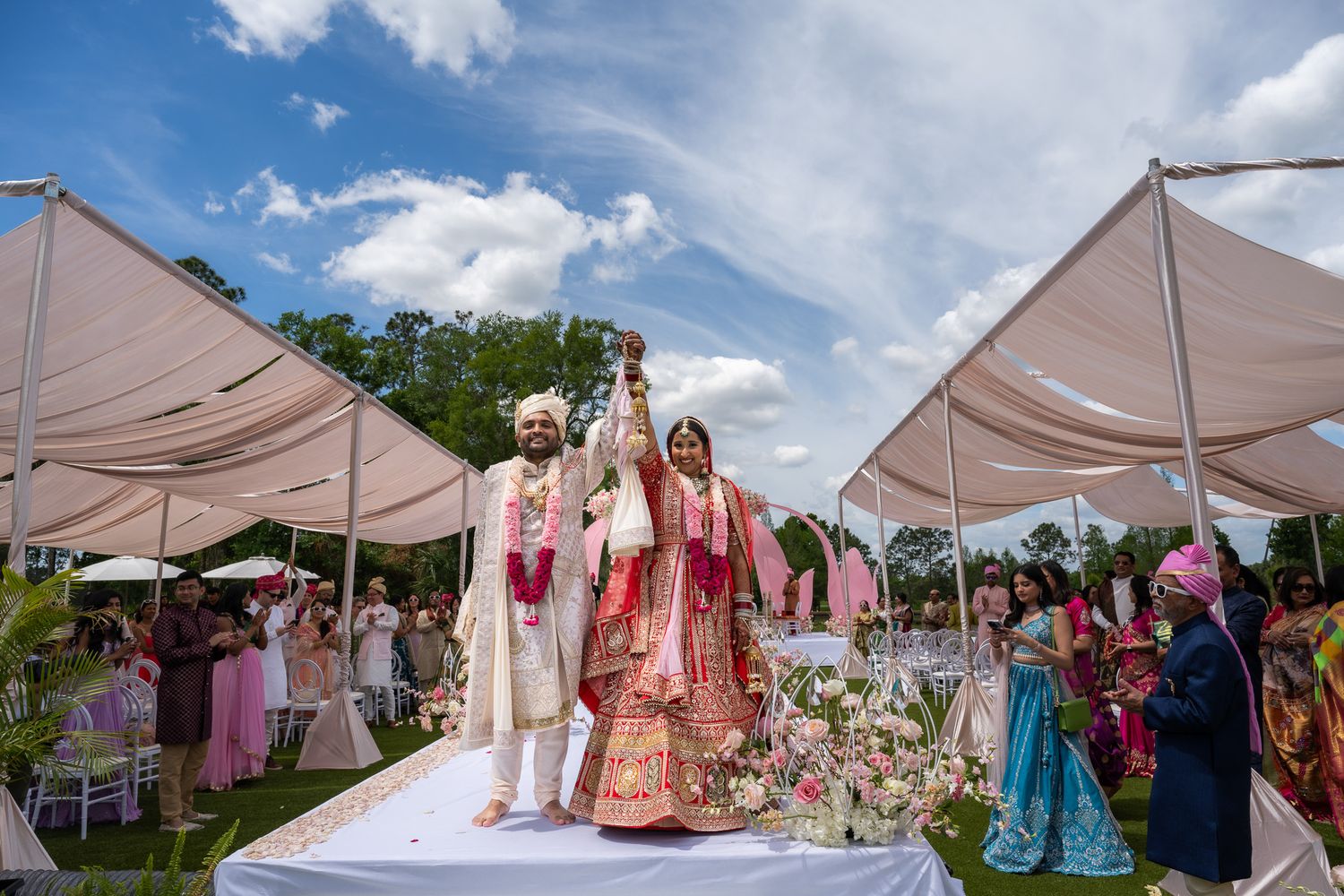 Outdoor Indian wedding ceremony under white canopy tents with floral decorations and gathered guests.