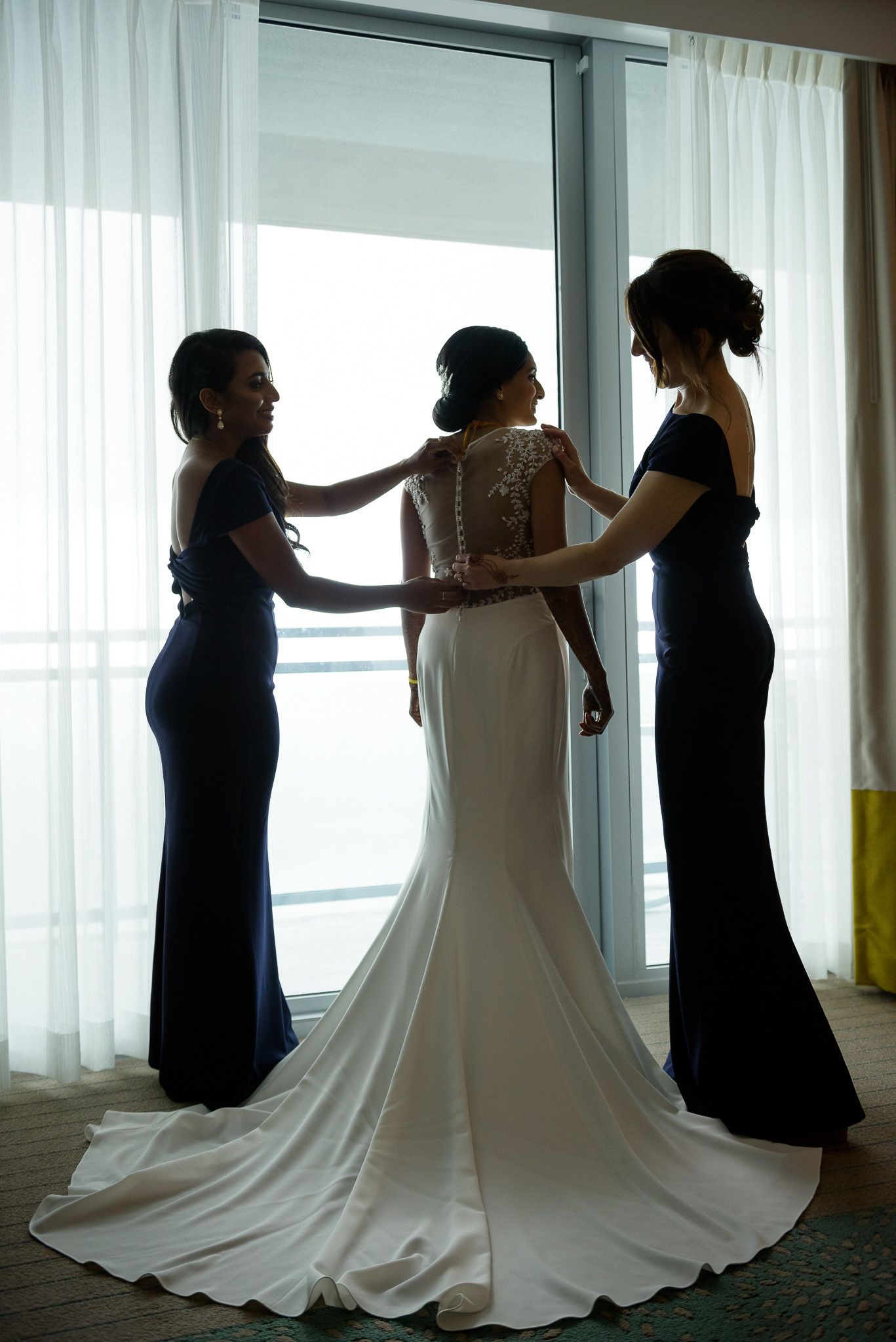 Three silhouettes stand by a window as they prepare a bride in her wedding gown.