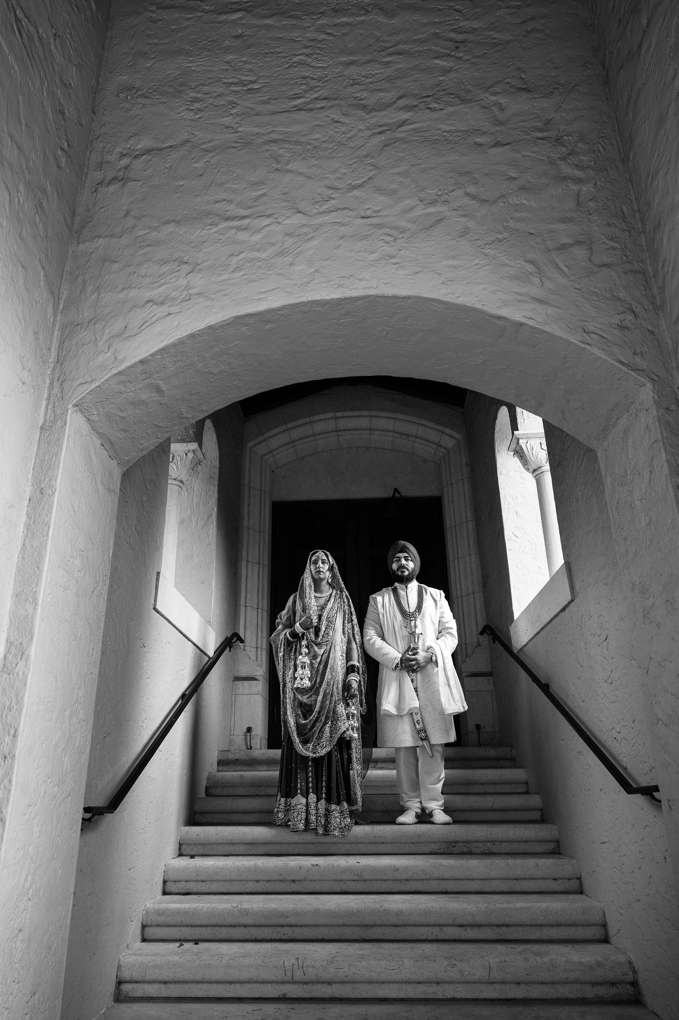 Black and white photo of two people in traditional dress standing in arched stone stairwell.