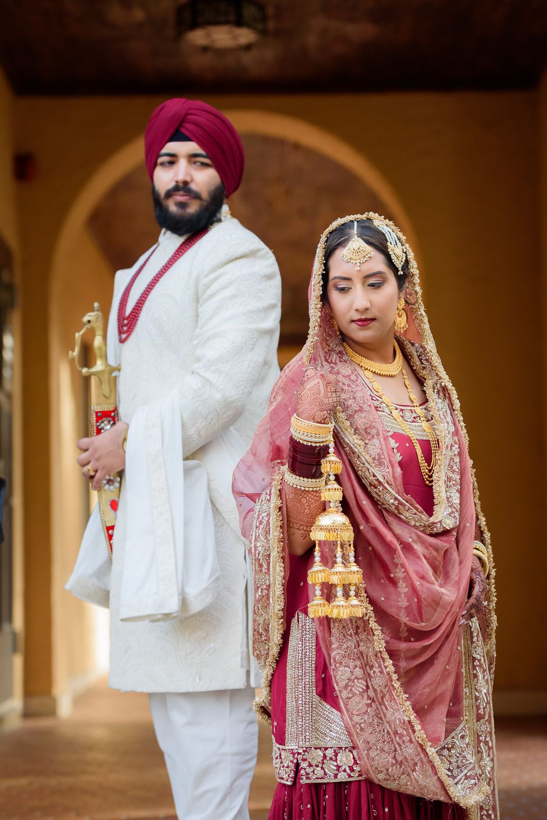 Traditional Punjabi wedding couple in elegant white and red attire under arched doorway.