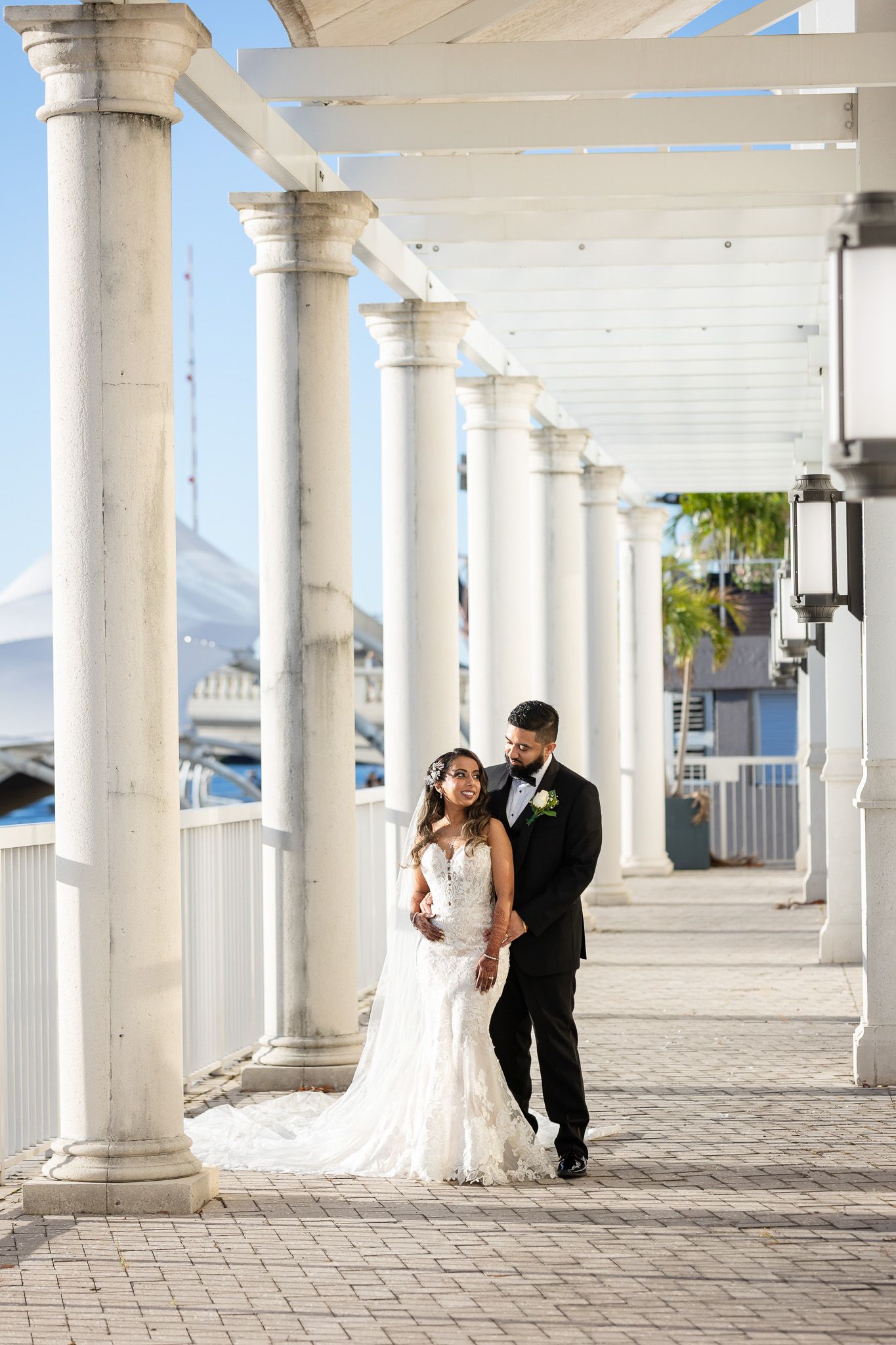 Wedding couple poses beneath white colonnade with beautiful lace gown trailing behind.