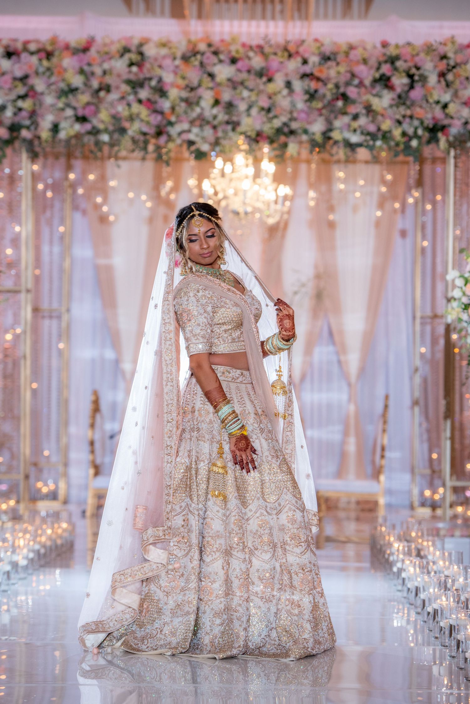 Bride in white and gold lehenga poses in front of floral backdrop with chandelier.