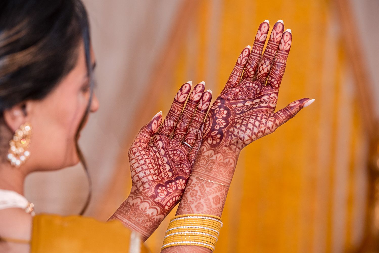 Detailed mehndi designs on raised hands with gold bangles against orange background.