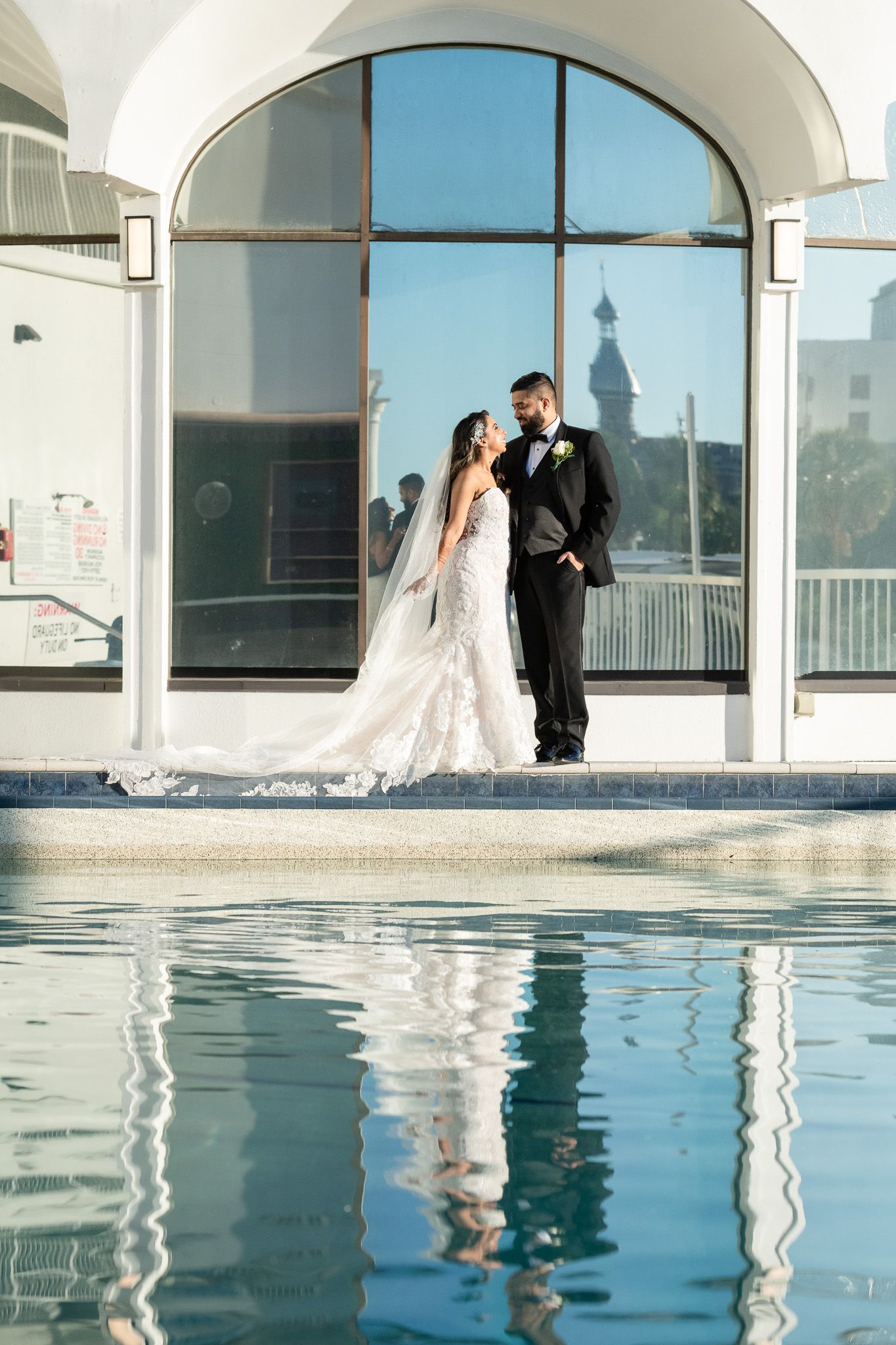 A wedding photo by a reflective pool with an arched architectural backdrop.