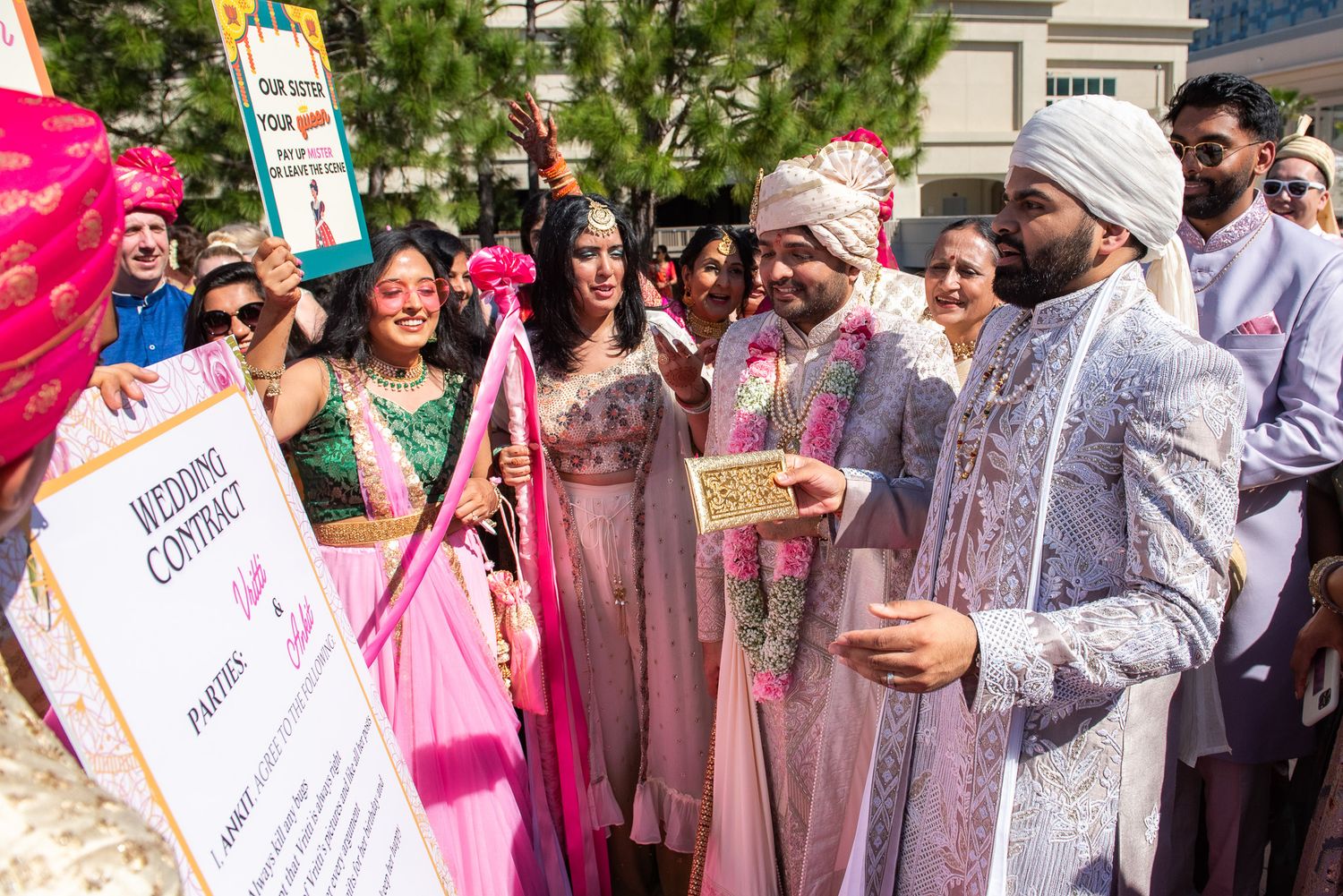 Celebratory group gathers outdoors in traditional South Asian wedding attire with colorful signs and decorations.