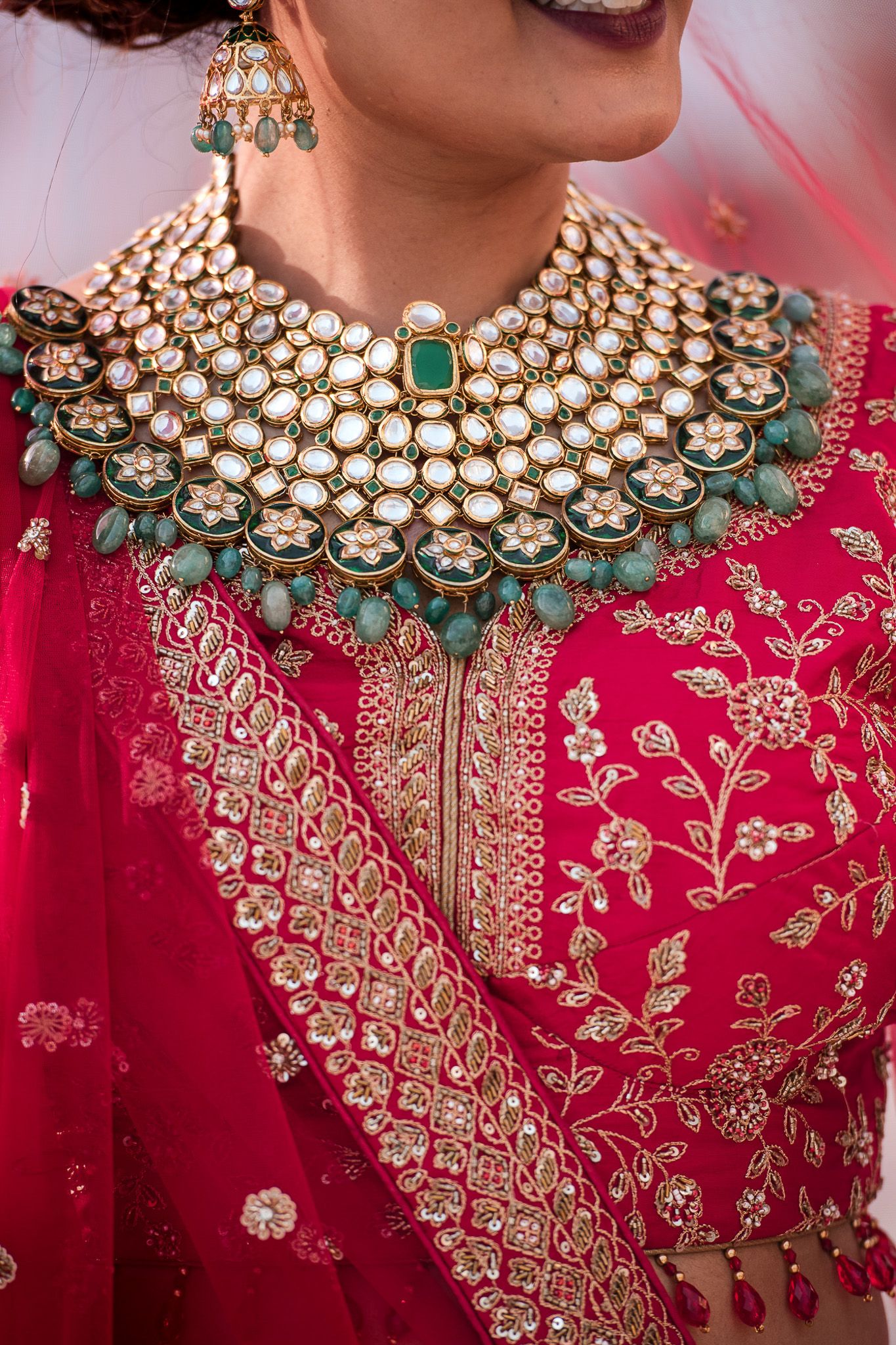 Close-up of ornate kundan necklace with emerald and pearl details on red embroidered fabric.