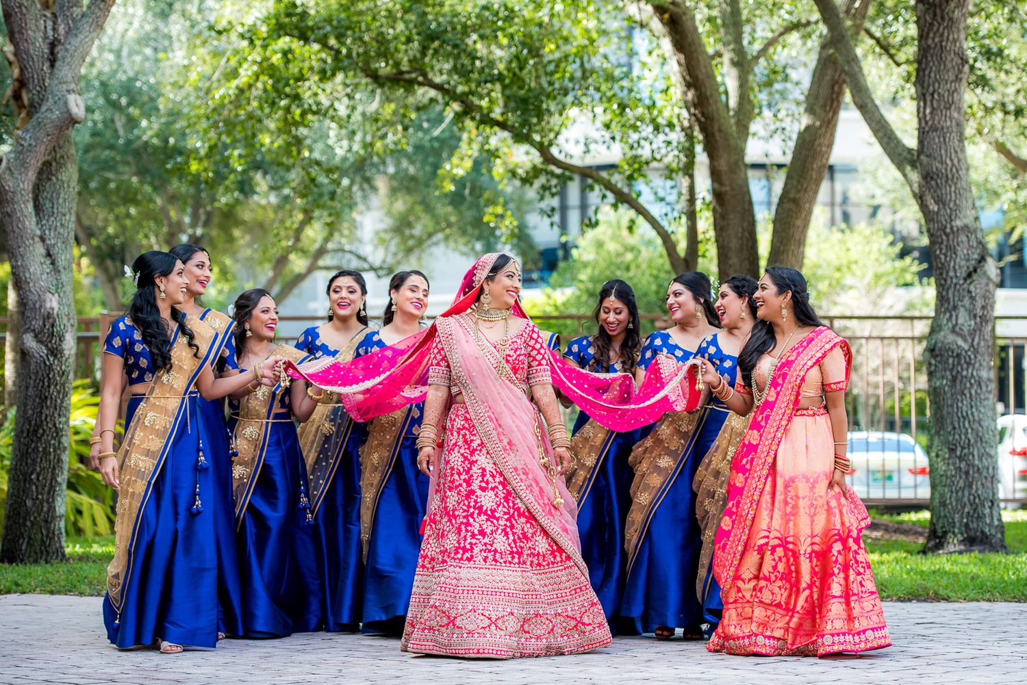 Bride in pink lehenga celebrates with bridesmaids in blue dresses during outdoor photoshoot.