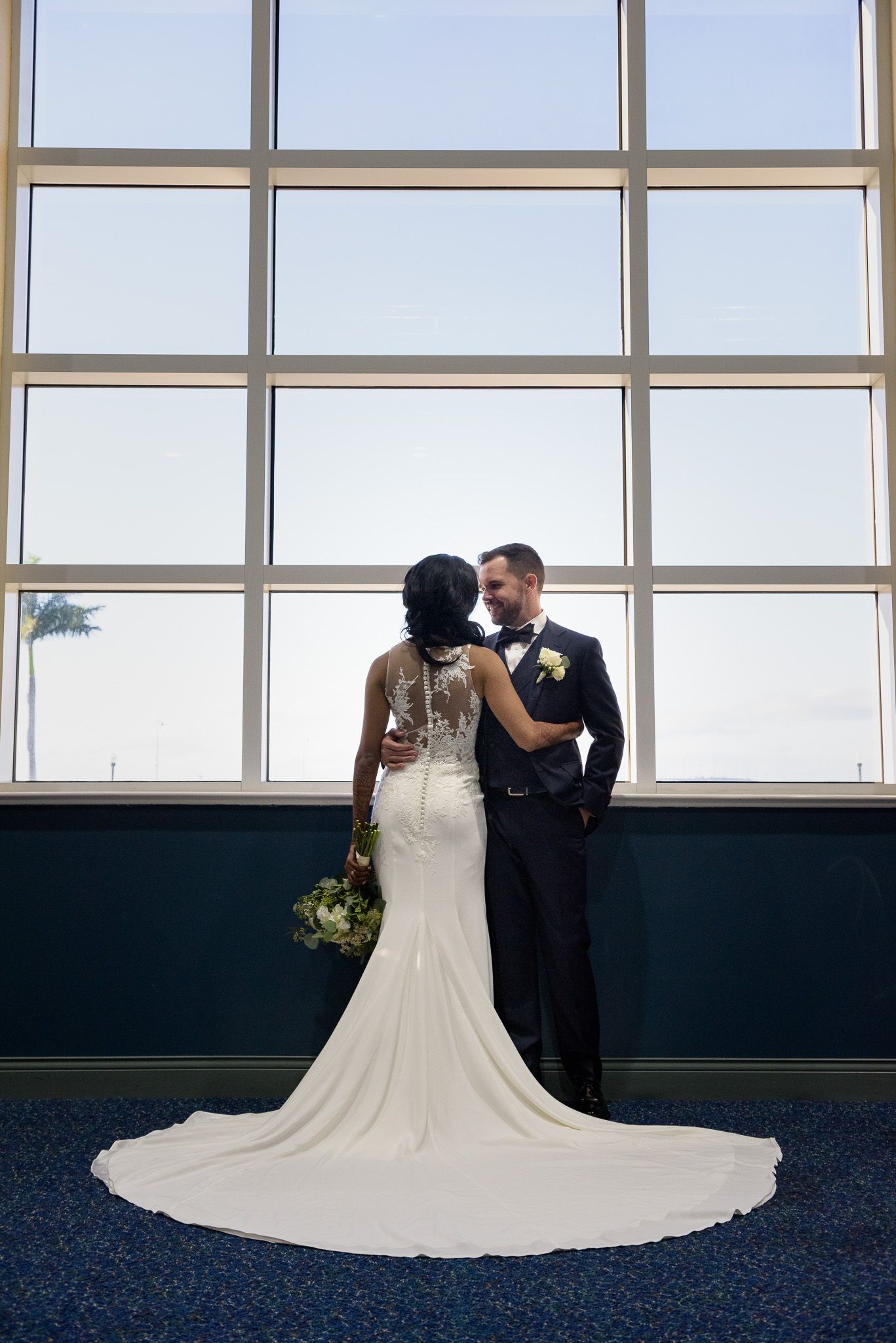 Bride and groom share intimate moment against large window backdrop with flowing white train.