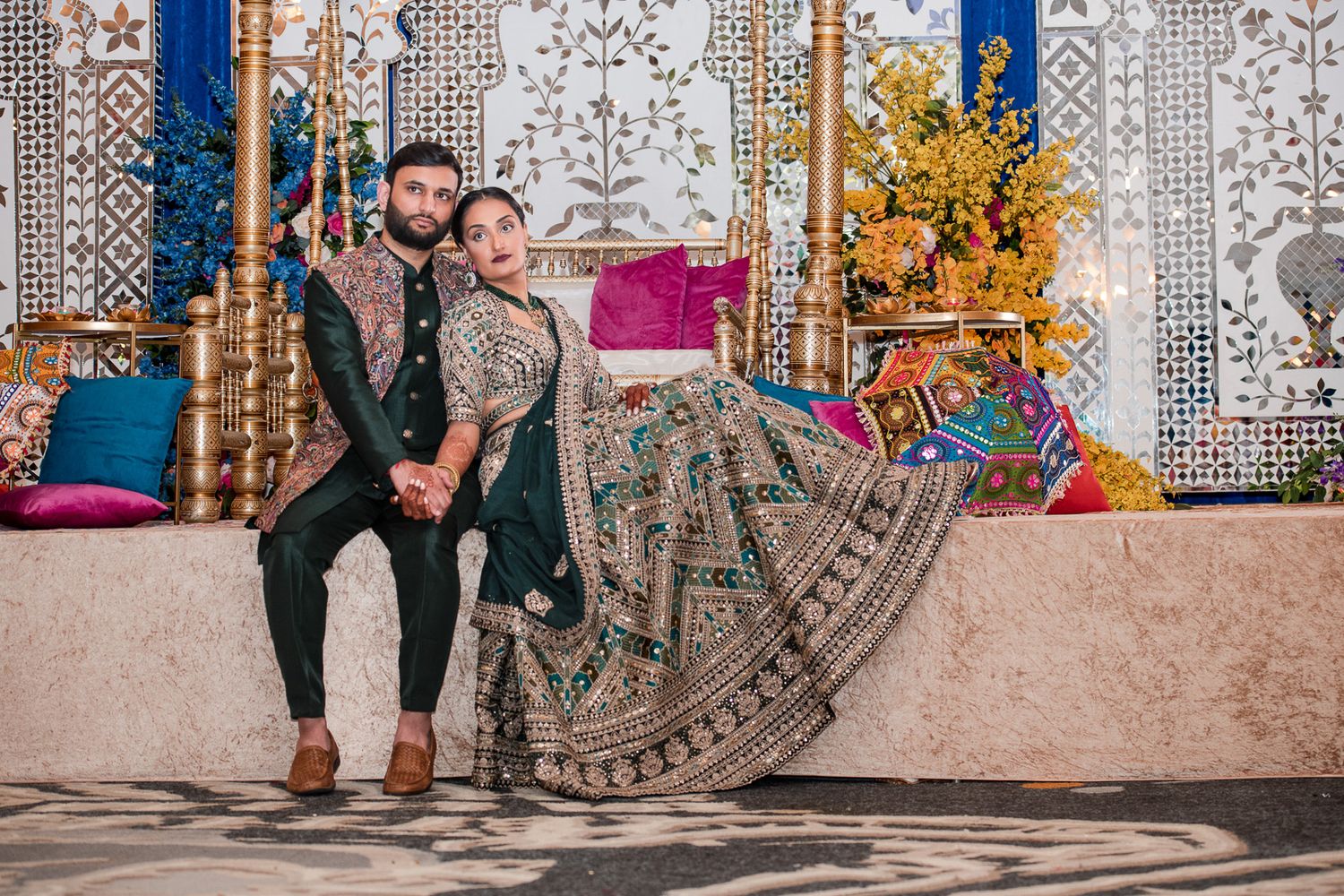 Couple in traditional South Asian wedding attire sitting together on an ornately decorated stage.